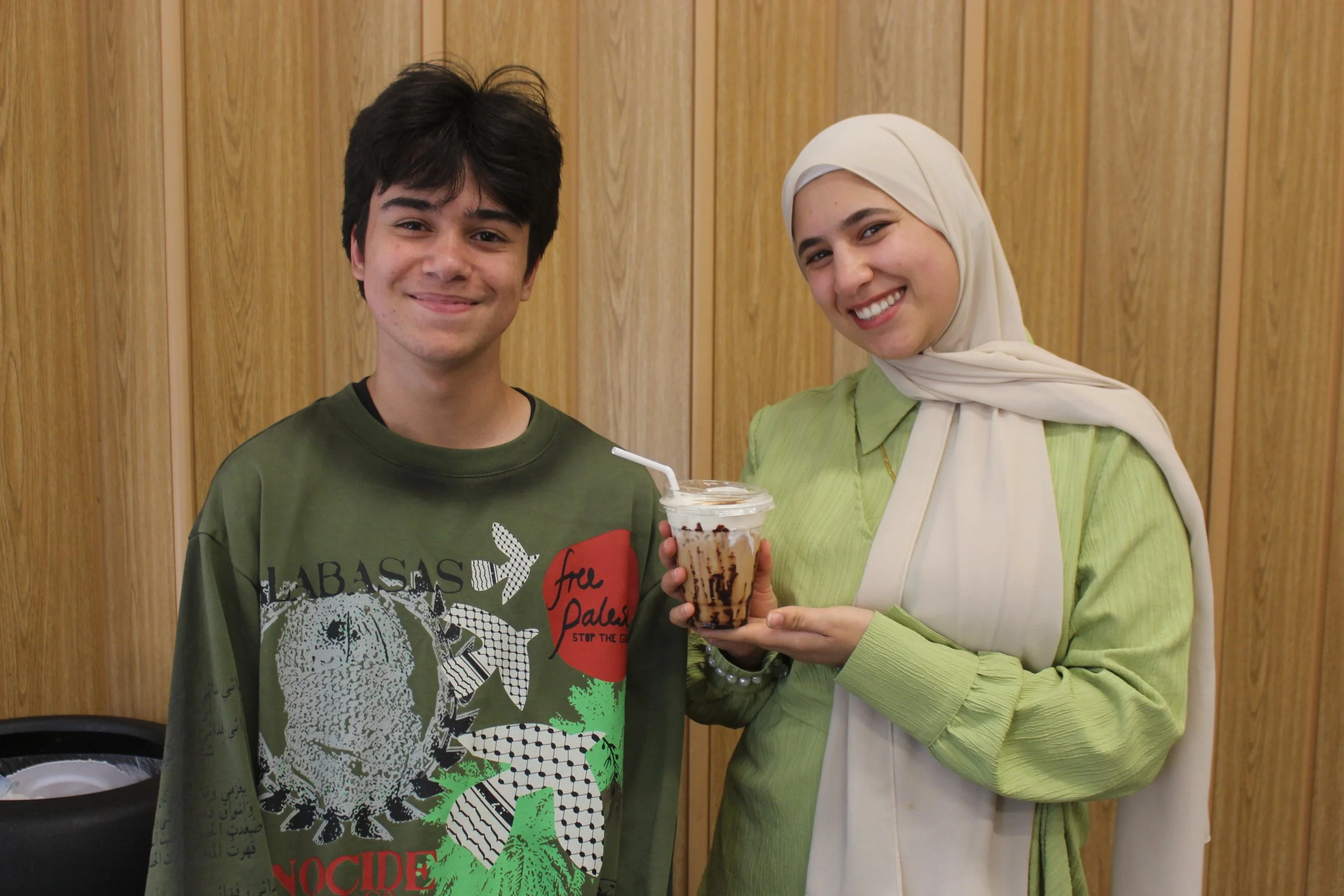 A young man and a young woman are standing together, smiling, with the woman holding a large iced beverage with whipped cream and chocolate syrup. They are in front of a wooden wall.