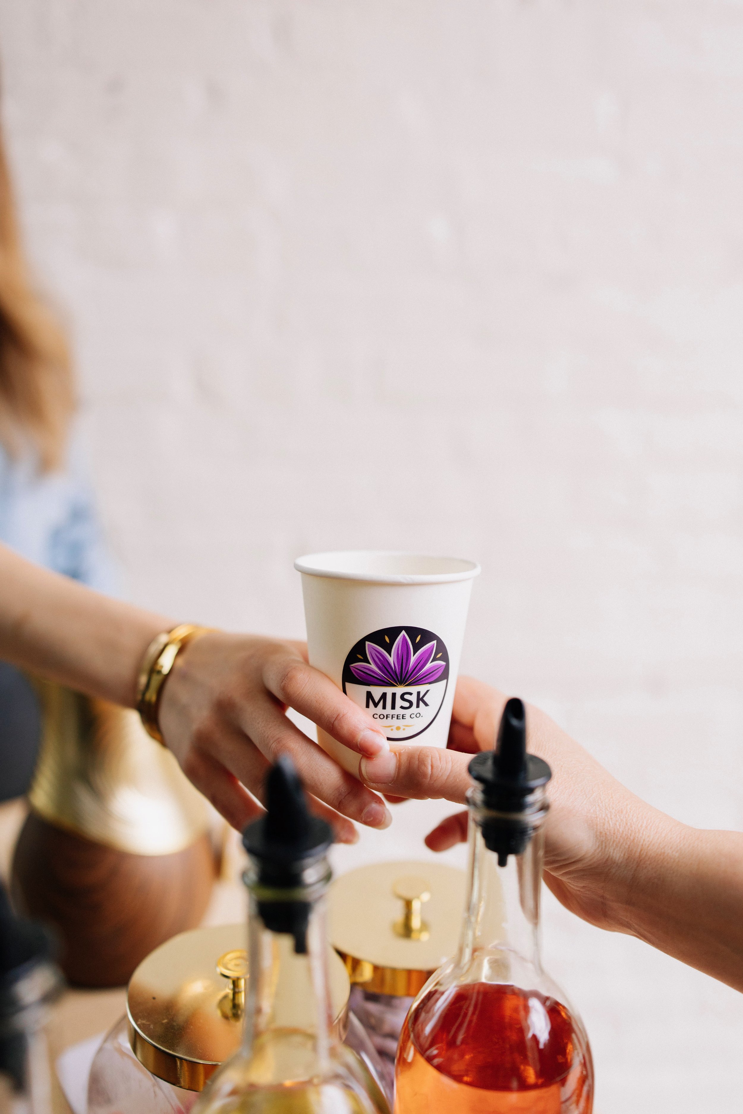 Close-up of a person holding a white cup with the logo "Misk Coffee Co." while filling it with a beverage. Decorative bottles are positioned on a circular tray in front of the person, with a white wall in the background.