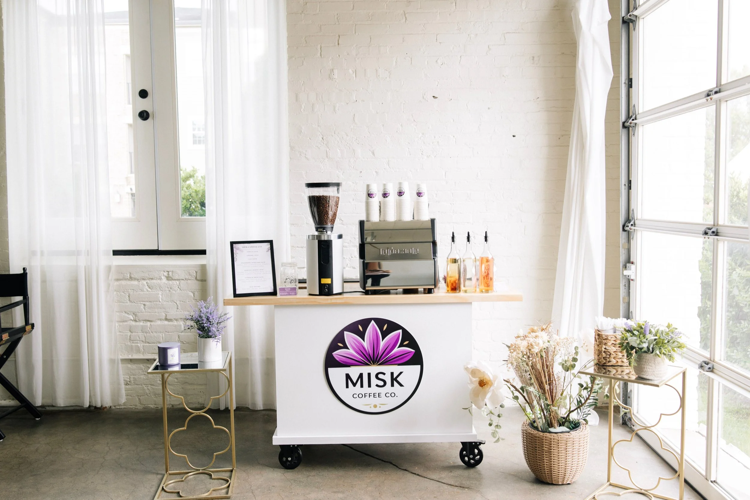 A coffee stand inside a bright space with white brick walls, large windows, and white curtains. The stand has a sign saying 'MISK Coffee Co.' and is equipped with a coffee grinder and cups. Decor includes flowers in a basket and gold-colored tables.