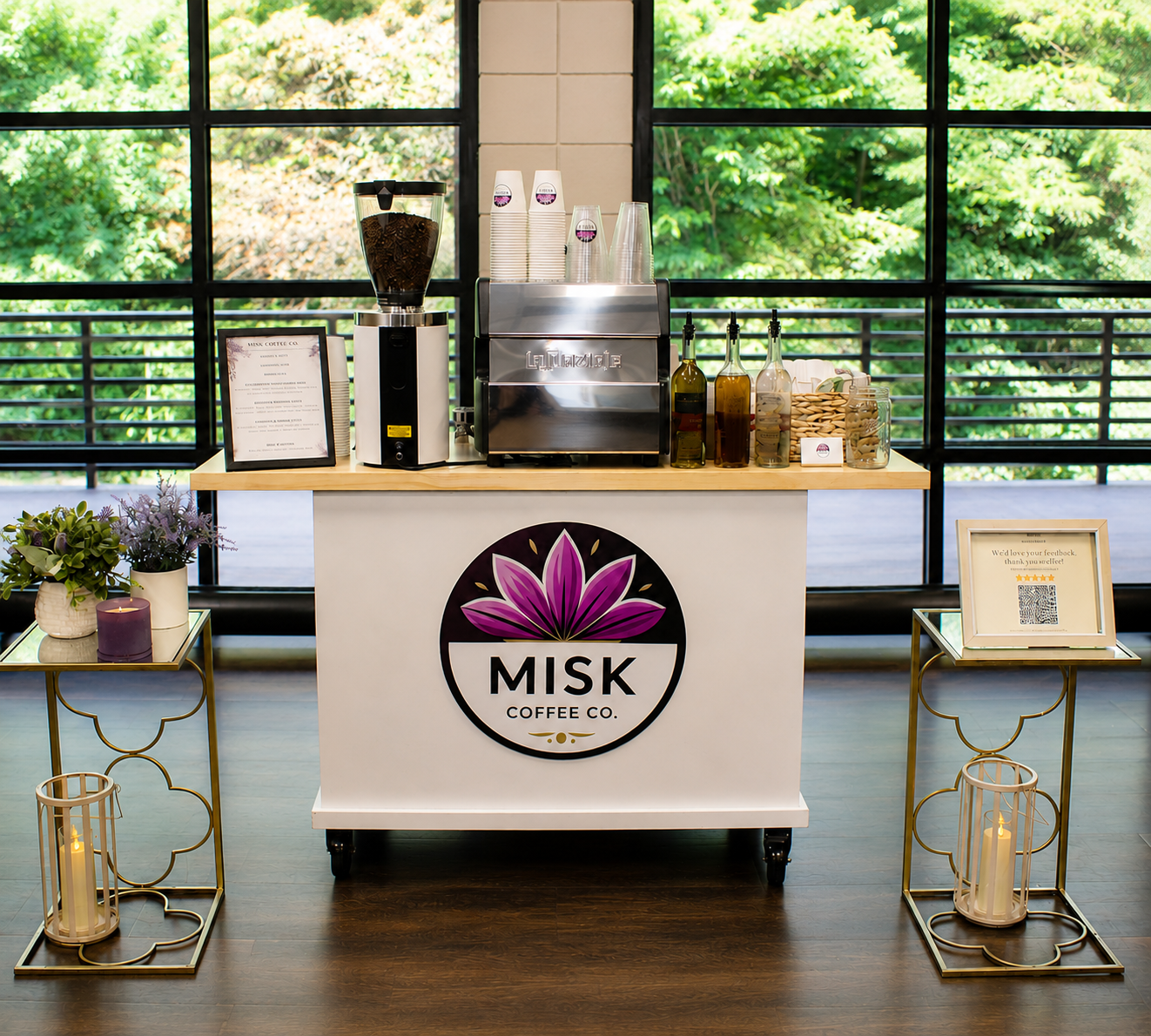 Coffee stand with a large logo of a purple lotus flower and the words 'Misk Coffee Co.' on the front, in front of large windows showing green trees outside. The stand has a coffee grinder, beverage dispensers, and bottles; beside it are two small tab