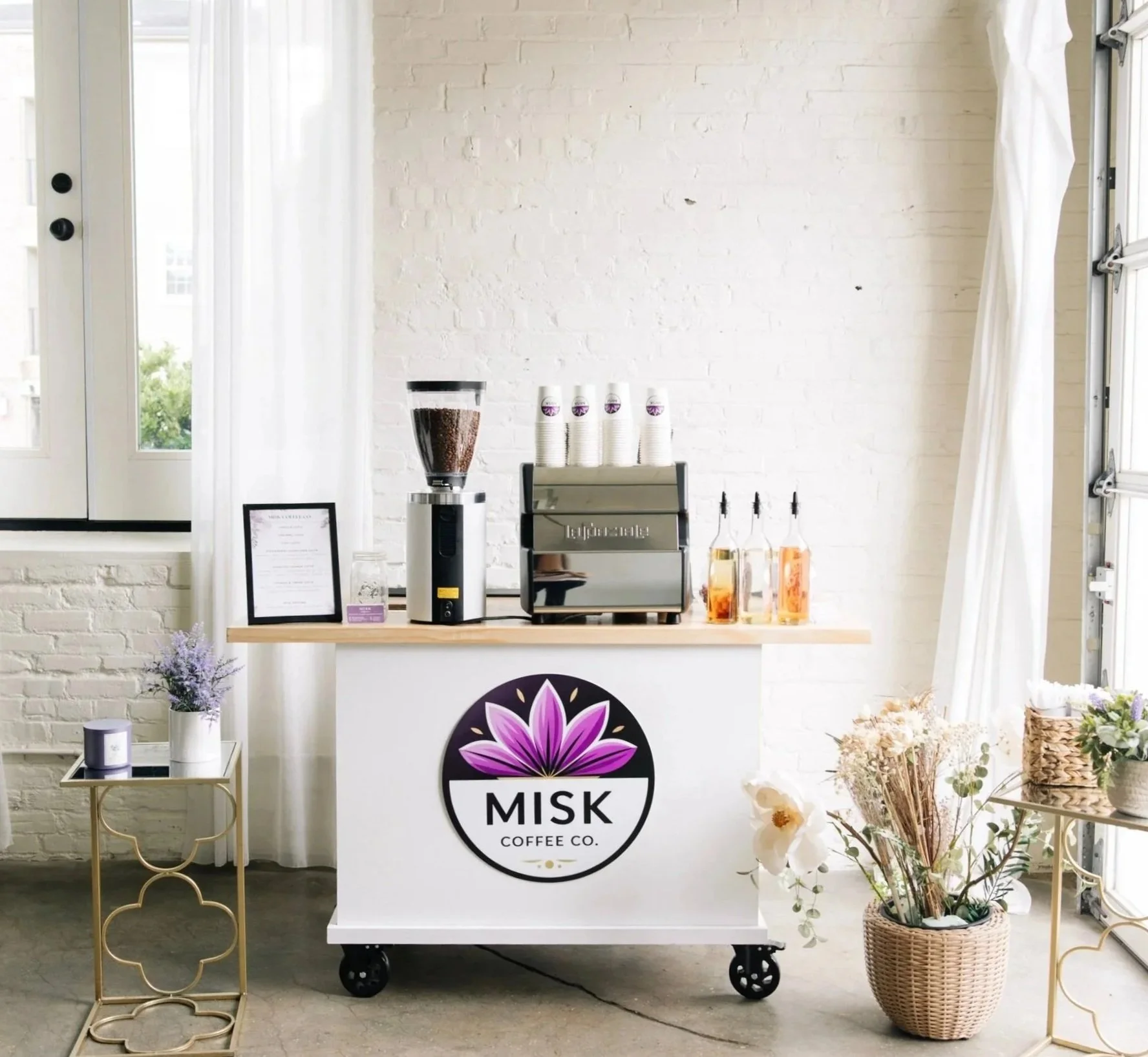 A coffee kiosk with the logo 'MISAK Coffee Co.' featuring a purple flower. Inside, a coffee grinder, cups, and bottles are displayed. Decor includes a small table with a candle and a potted plant, along with flowers in a woven basket.