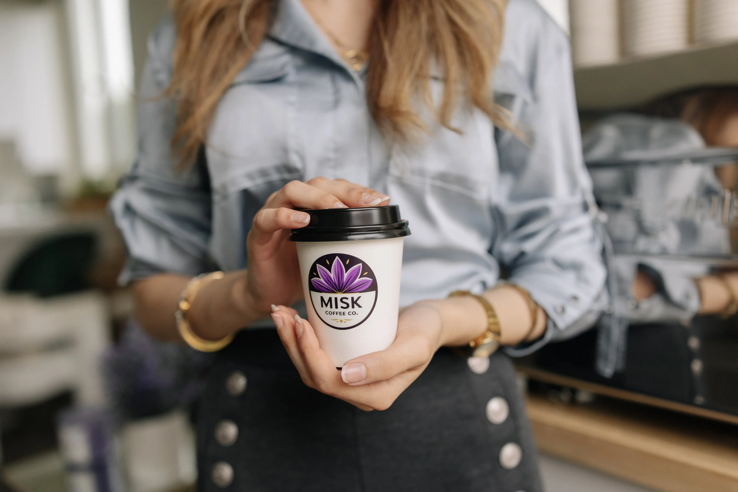 Person holding a takeout coffee cup with a logo that says 'MIK Coffee Co.' logo features a purple flower. The woman is wearing a light-colored denim shirt and gold jewelry, standing in what appears to be a coffee shop or cafe.