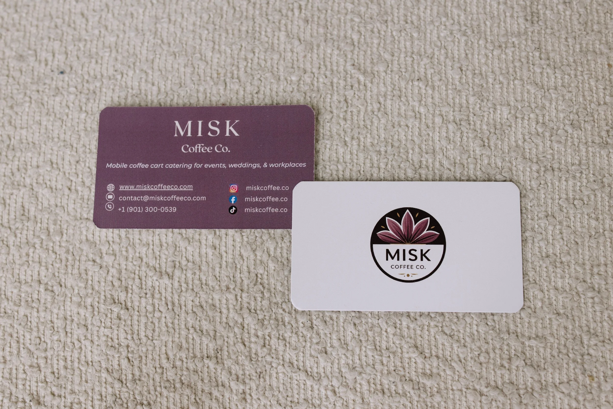 Close-up of two business cards on a beige textured surface. The card on the left is purple with white text that reads 'MISK Coffee Co.', and includes contact information and social media icons. The card on the right is white with a logo featuring a f