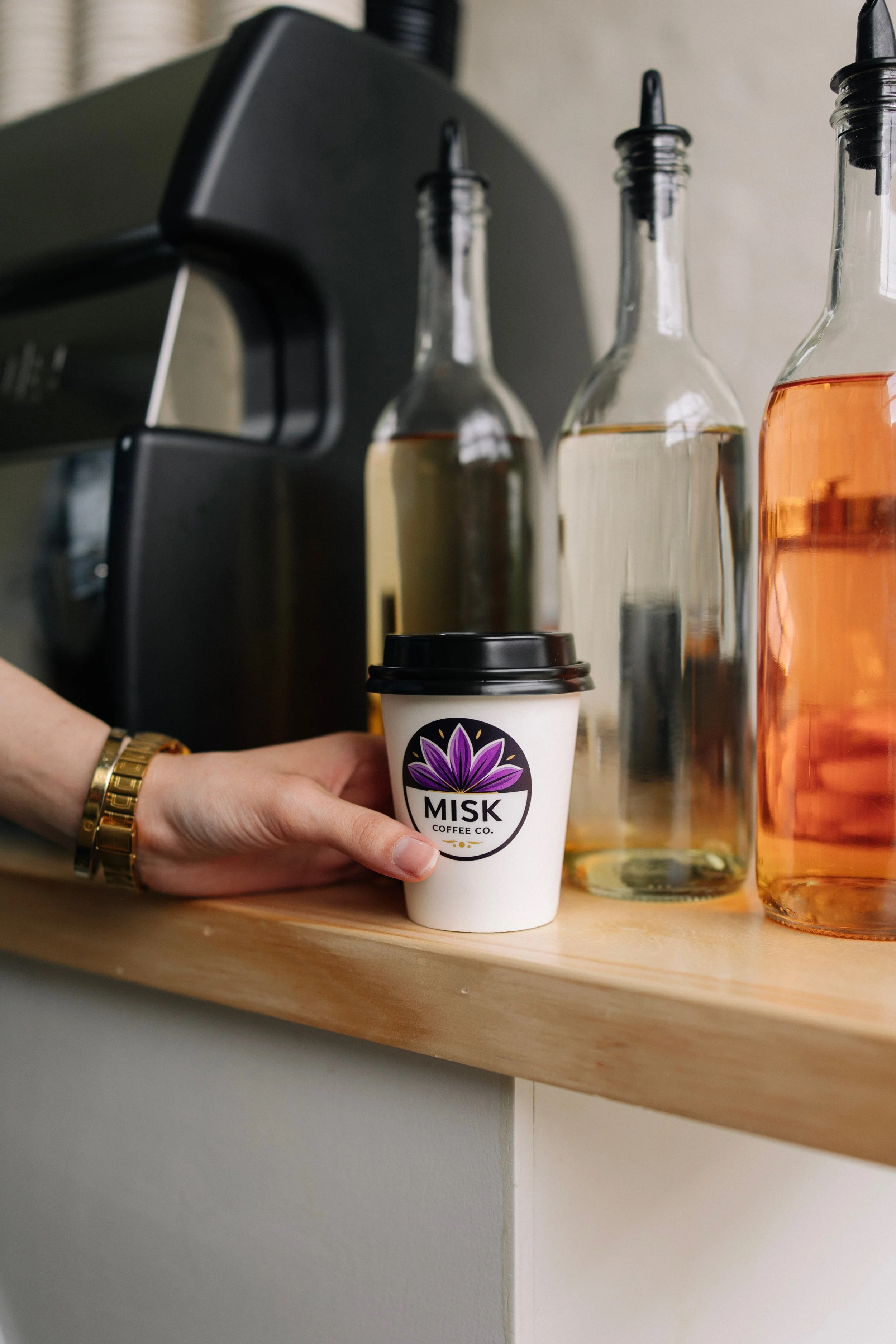 A hand holding a paper coffee cup with the logo of Misk Coffee Co. on it, with bottles of liquid in the background.