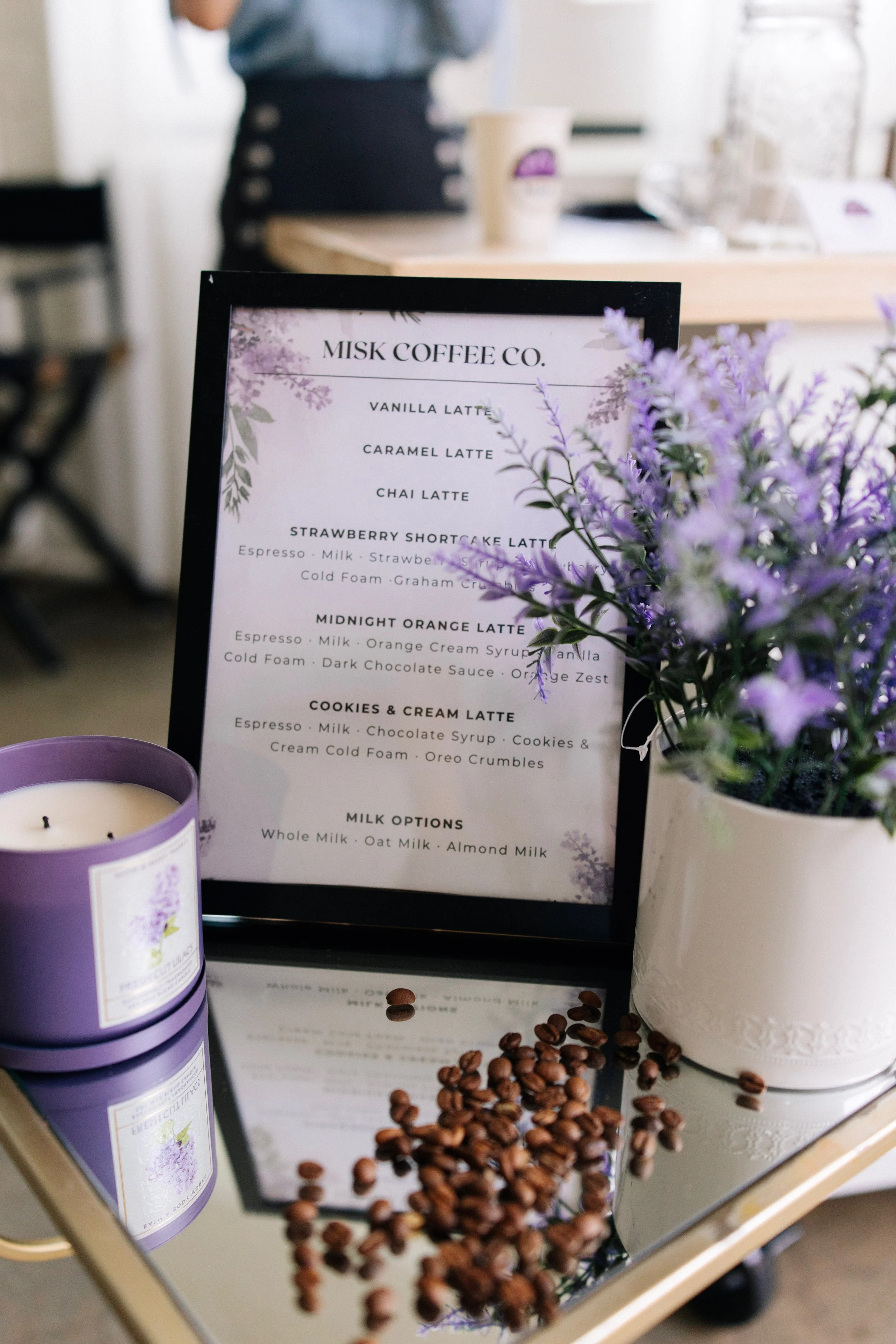 Close-up of a coffee shop menu with purple flowers and a candle in the foreground, and a person behind the counter in the background.