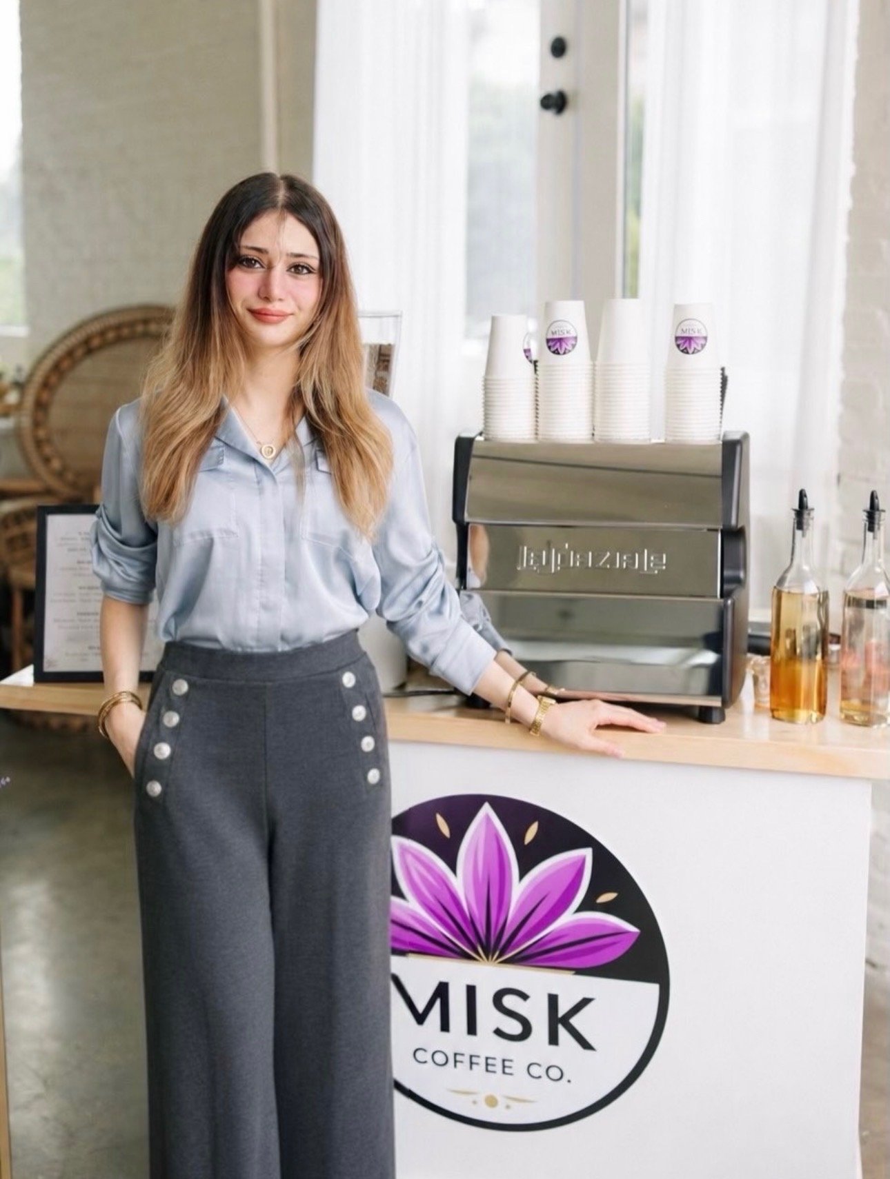 A woman standing behind a coffee stand with a logo that says 'MISK Coffee Co.' The stand has disposable cups, glass bottles with liquids, and a coffee machine. The woman is wearing a light gray shirt with rolled-up sleeves and dark gray high-waisted pants with large buttons.