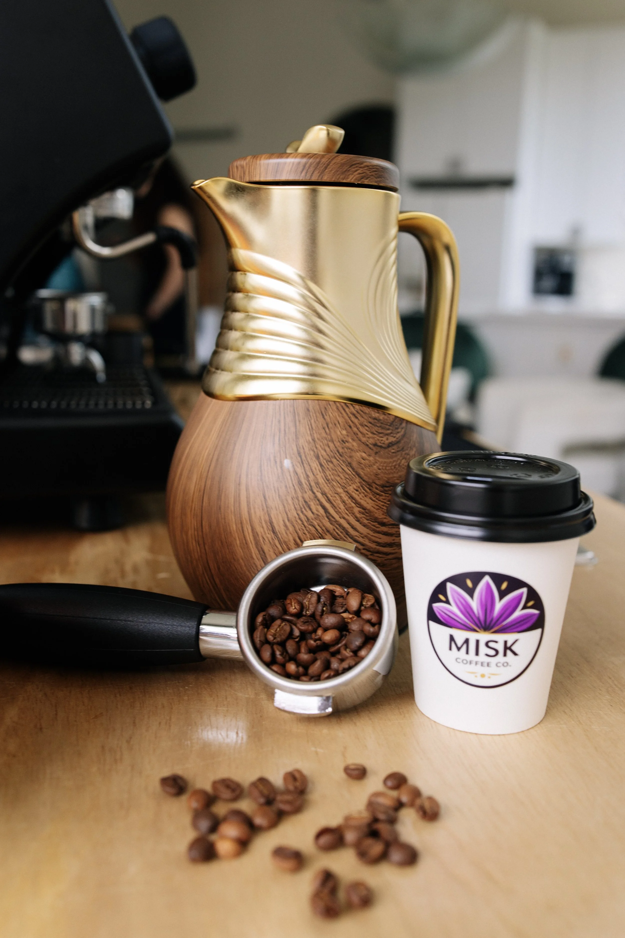 A coffee setup featuring a golden and wooden teapot with a spiral design, a small bowl of coffee beans, a to-go coffee cup labeled "Misk Coffee Co." with a purple and pink flower logo, a portafilter with coffee beans, and scattered coffee beans on a wooden surface, with a coffee machine and kitchen in the background.