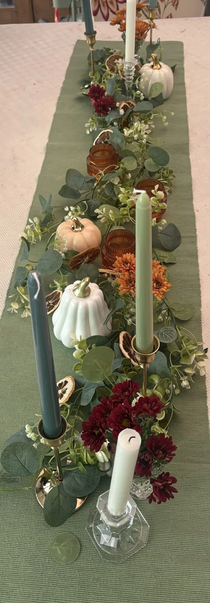 Autumn Harvest table styling with sage green runner, eucalyptus garland, amber glass votives, pastel pumpkins, dried orange slices, and mixed taper candles in soft green, blue, and ivory tones.