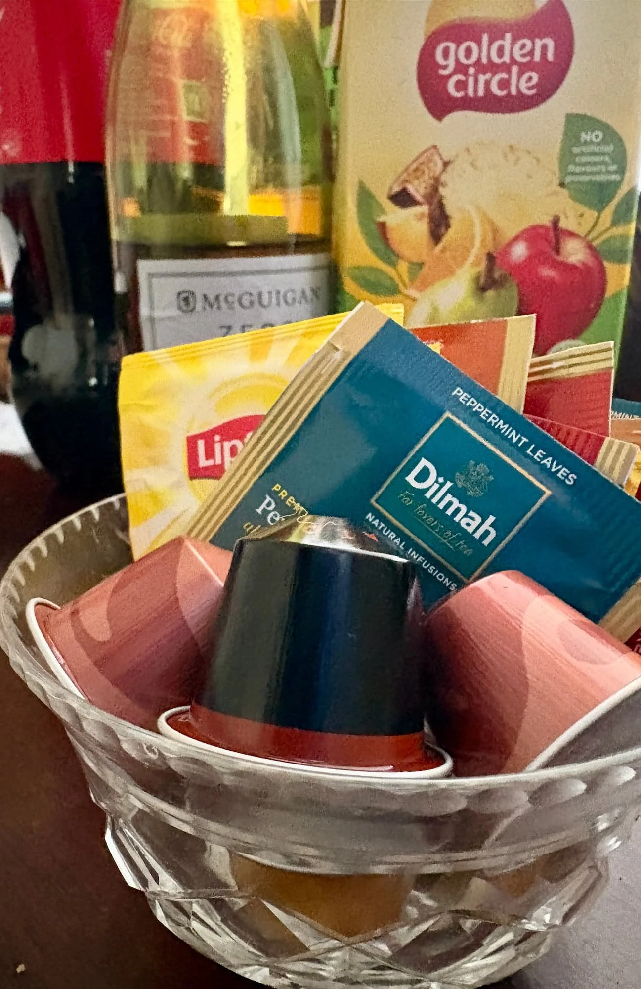 small glass bowl filled with coffee pods and tea bags,a carton of fruit juce and a bottle of wine are in the background