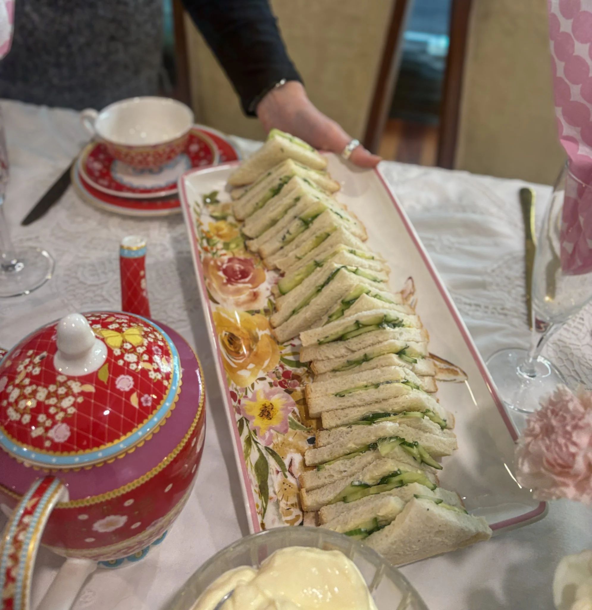 Plate of triangle shaped sandwiches filled with pale green cucumbers are placed on a table, A bright red teapot is in the foreground