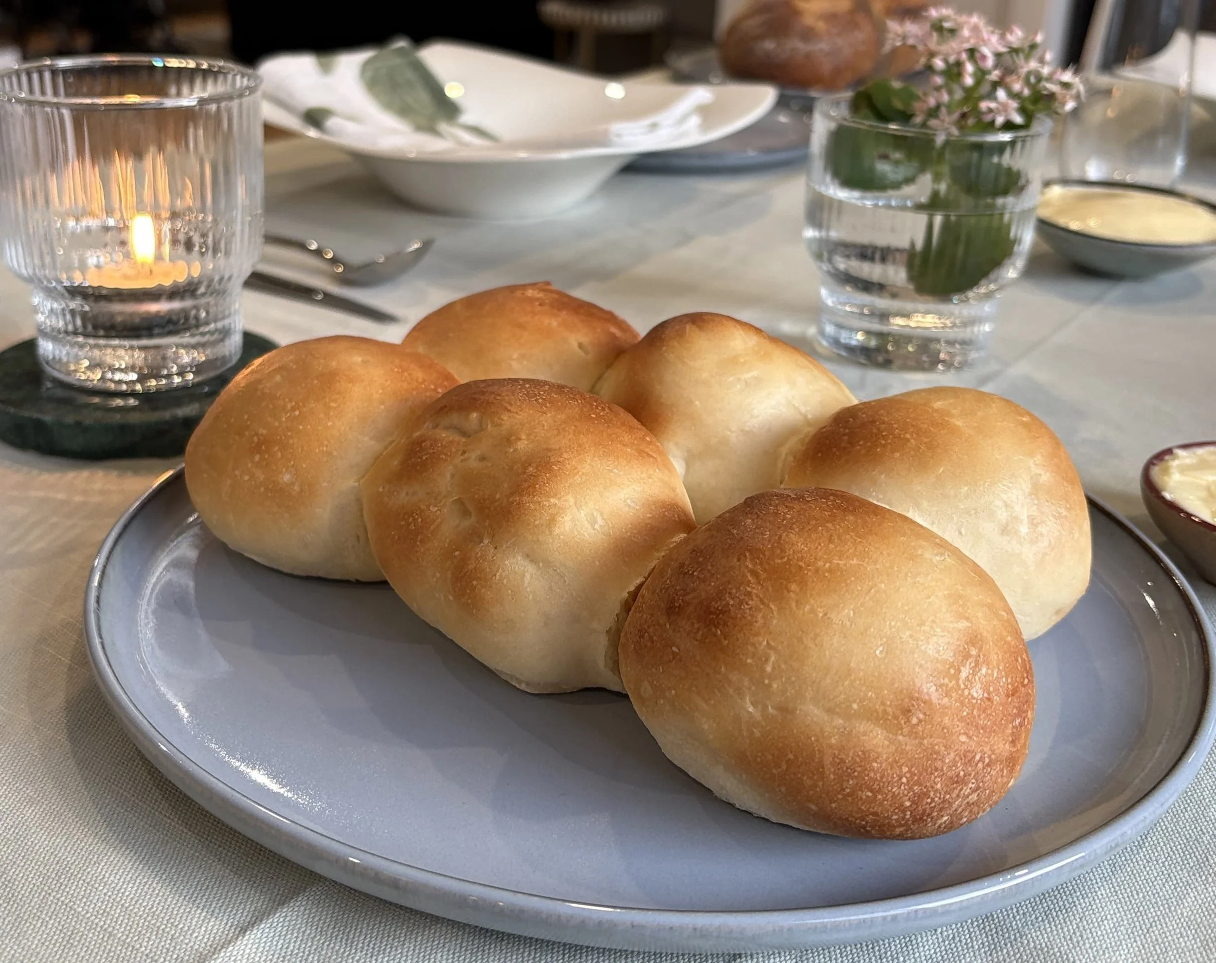 6 homemade bread rolls on a plate. In the background is a candle, small pink flowers in a vase and a bowl with a green patterned napkin laid over the top