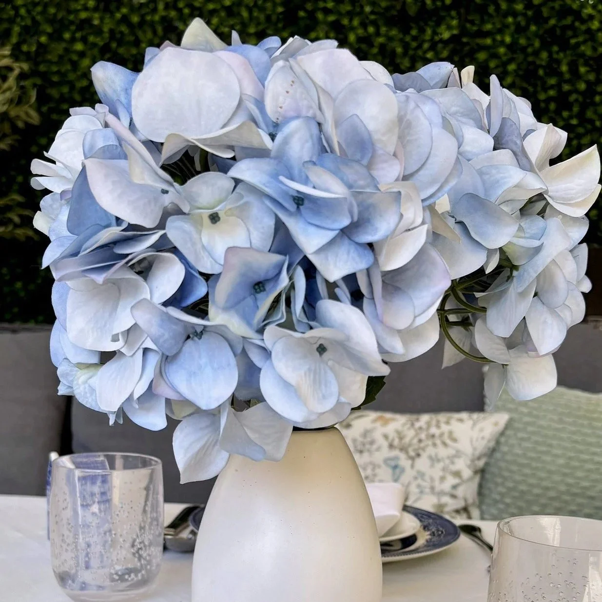 Outdoor garden table setting featuring a large blue hydrangea centerpiece in a white vase, blue and white porcelain plates, silver cutlery, and bubble-textured glassware on a white tablecloth.