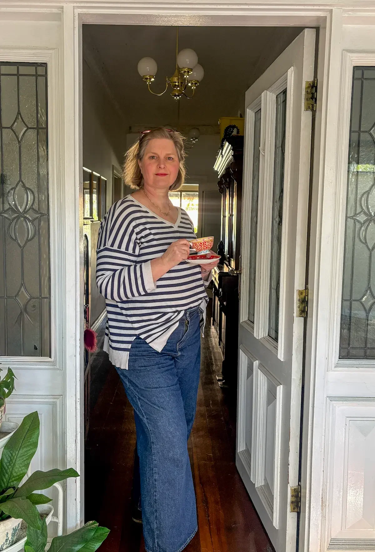 A woman wearing a blue and white striped top holds a red and white teacup and saucer. She is staning in the open doorway of a house with leadlight windows on either side of her