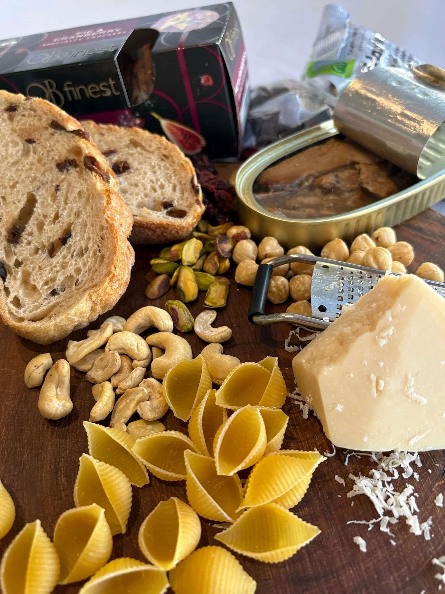 pasta shells, slices of bread, a chunk of cheese and an opened tin of sardines rest on a wooden platter