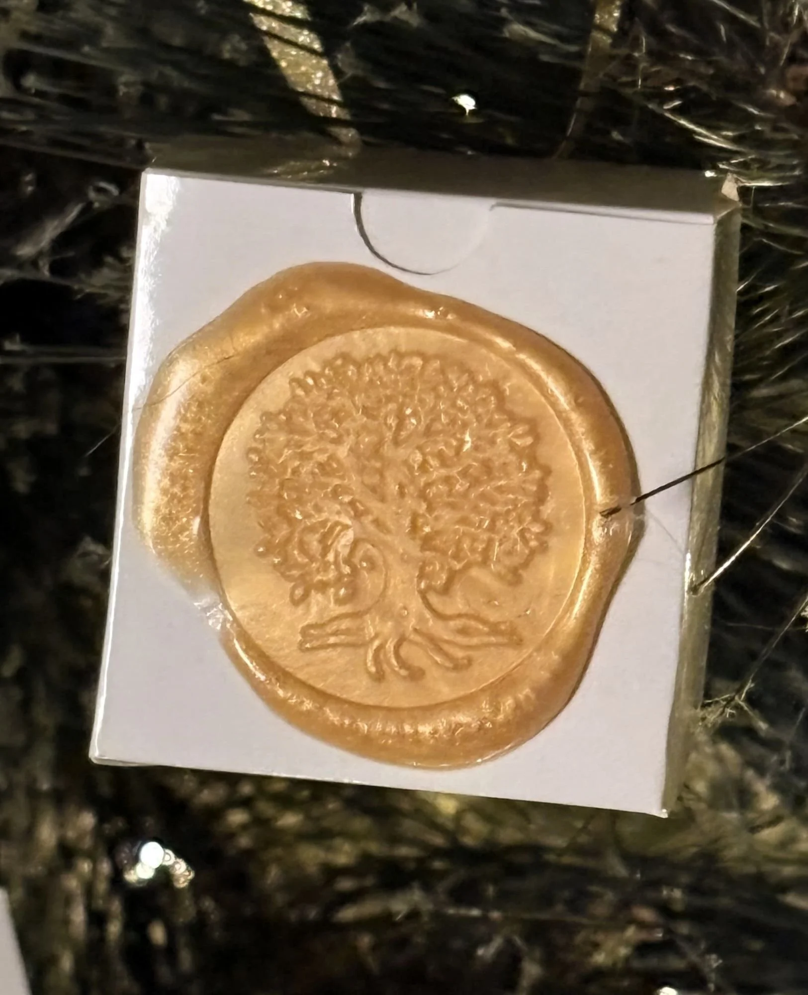 A small white gift box with a gold wax seal on the front hangs from the branches of a Christmas tree