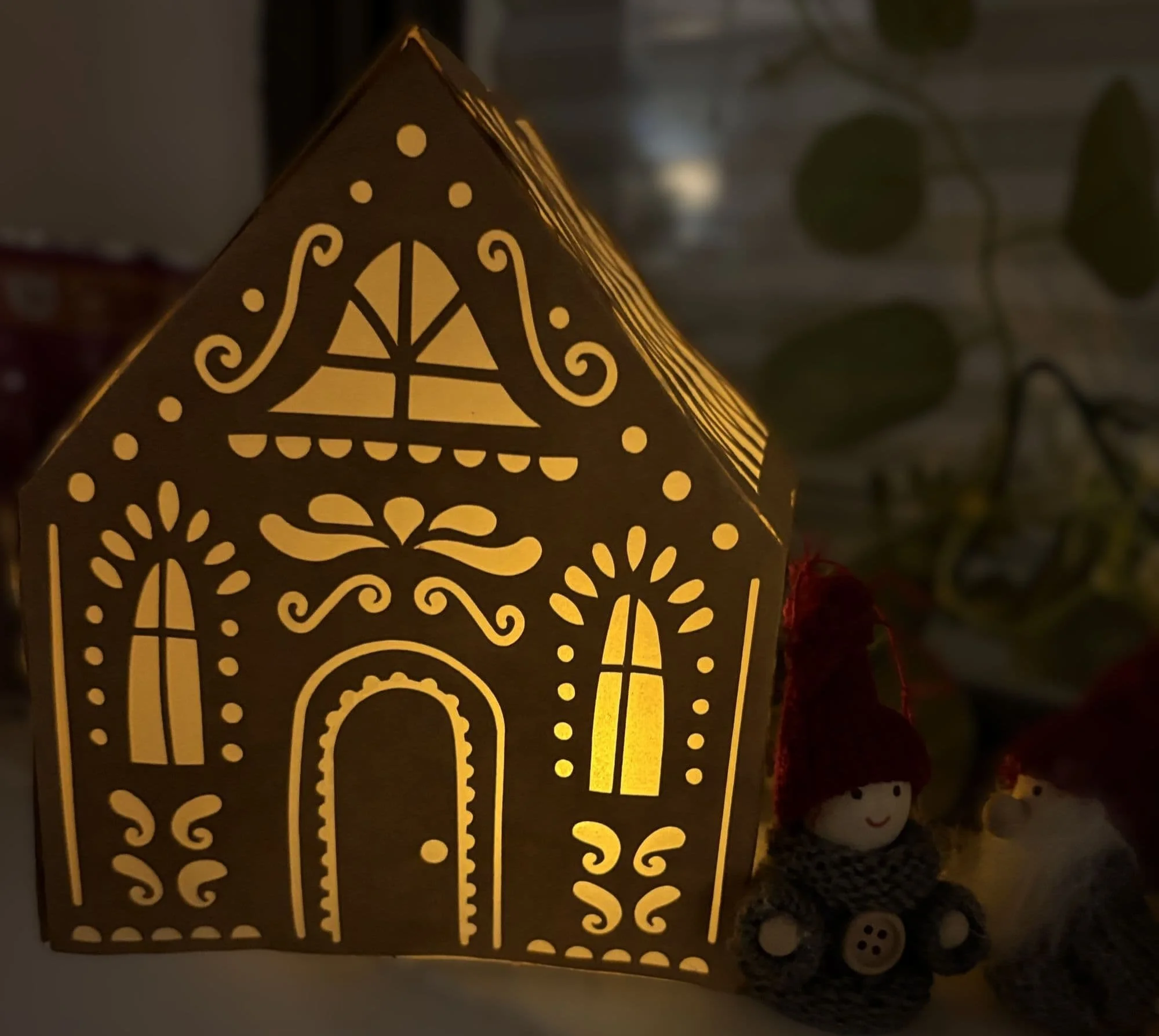 Gingerbread style house made of brown paper lit with an LED candle glows in dim light. Two stylised figurines in woolly hats and coats are placed to the right of the house. Green leaves are in the background.