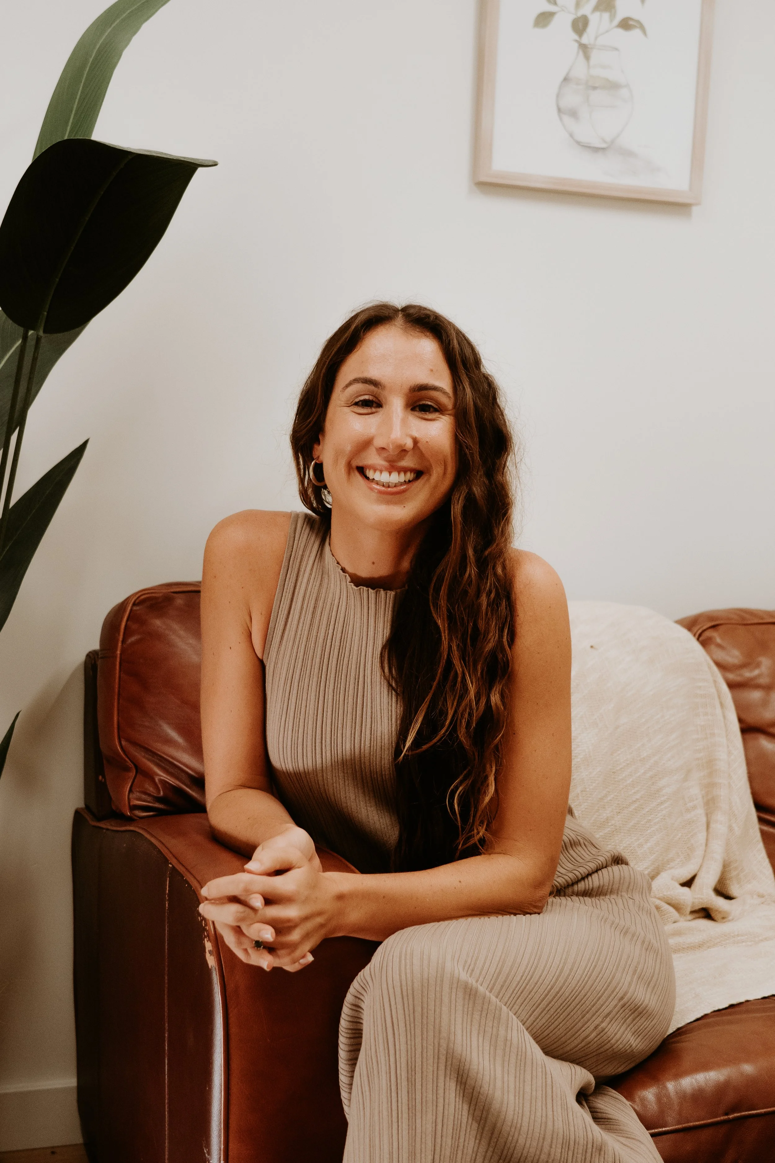 A woman with long, curly brown hair and a radiant smile, sitting on a brown leather sofa in a cozy room with white walls, decorated with framed artwork and a large green plant.