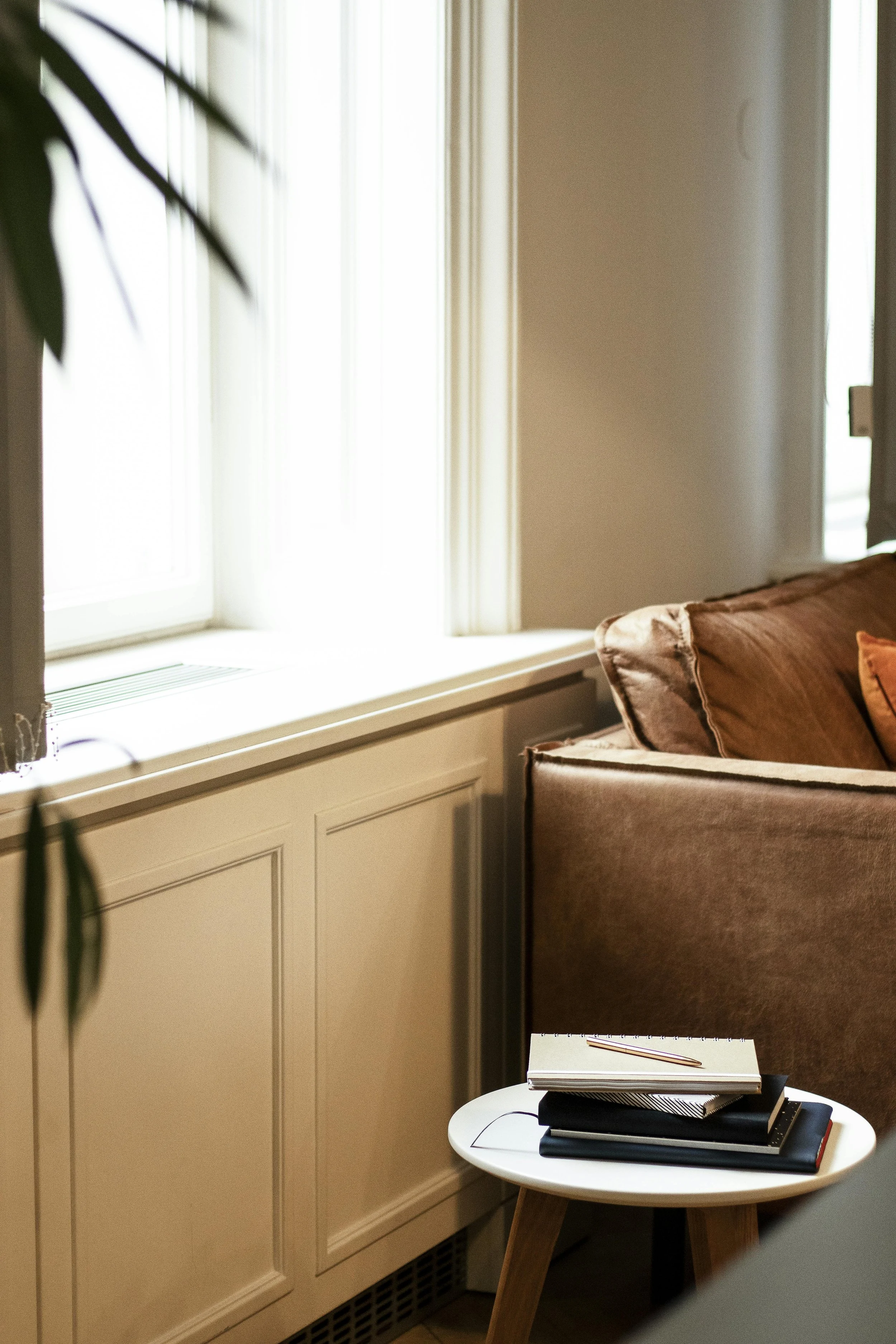 A cozy corner of a living room featuring a beige sofa with brown cushions, a small white side table with notebooks and a pencil, and a large window with curtains.