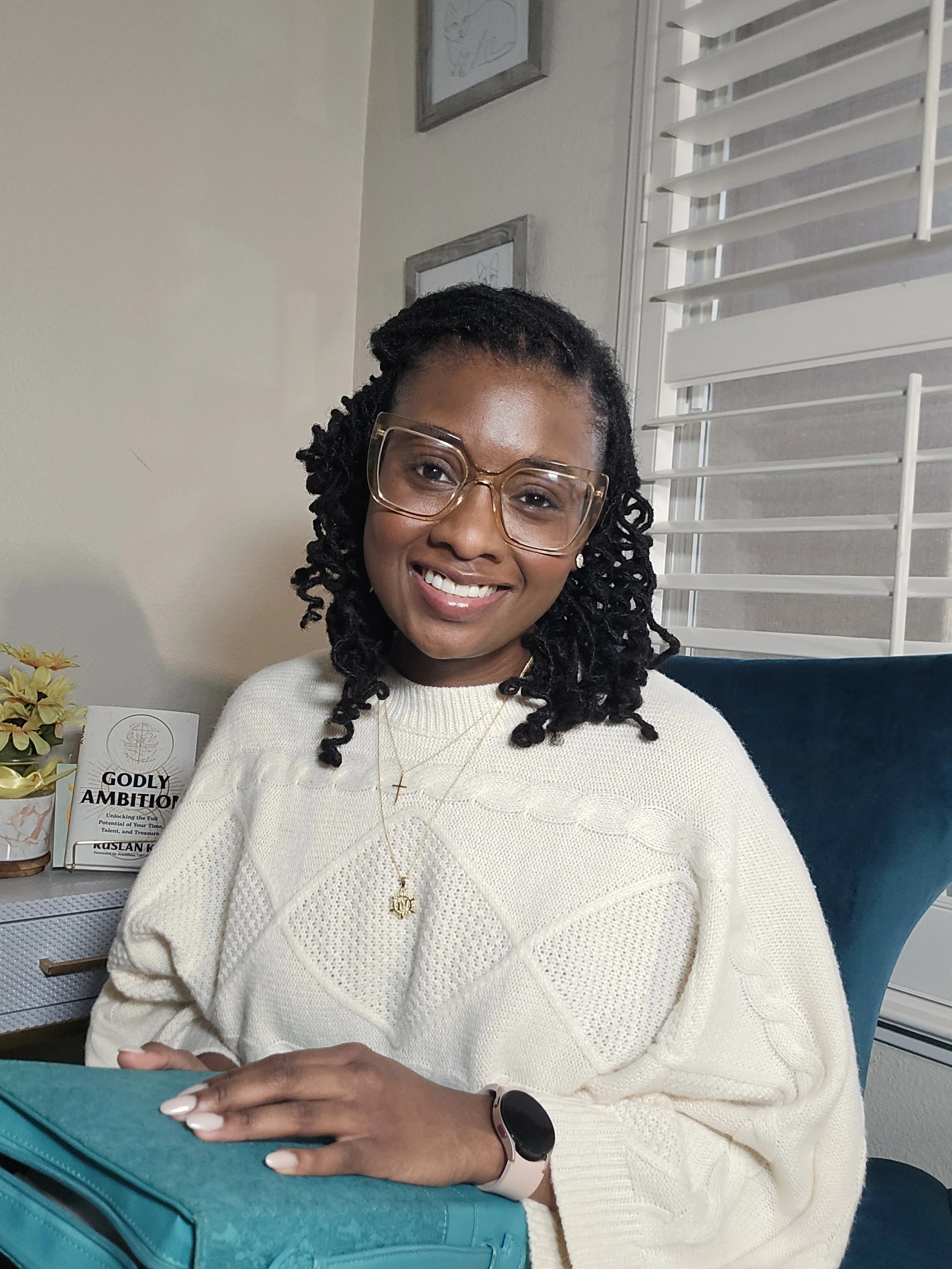 A woman smiling, wearing glasses, a white sweater, and gold necklaces, sitting at a desk with a teal bag in front of her, in a room with white blinds and framed artwork on the wall.