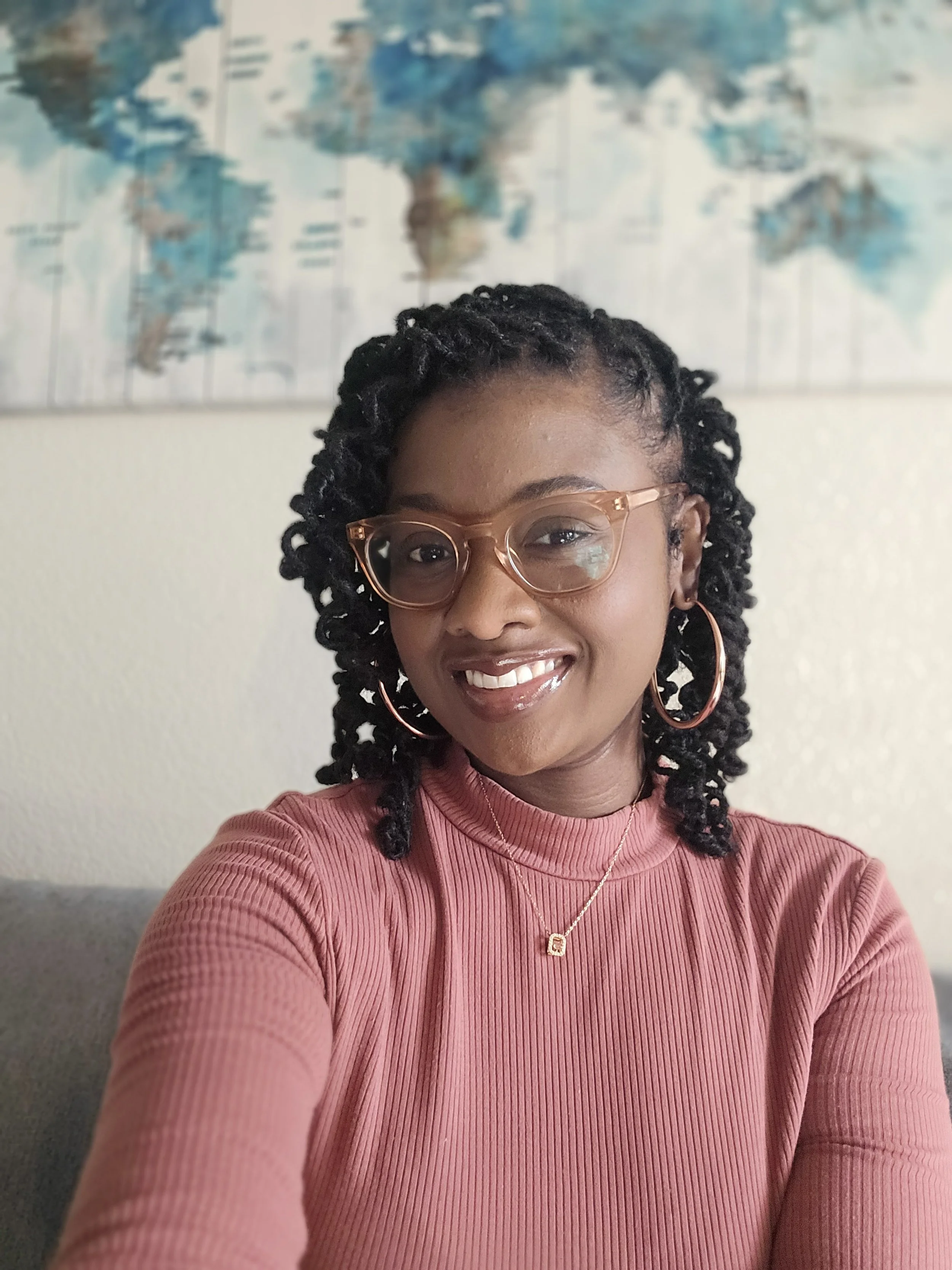 A woman with glasses and hoop earrings taking a selfie, smiling, with a world map in the background.