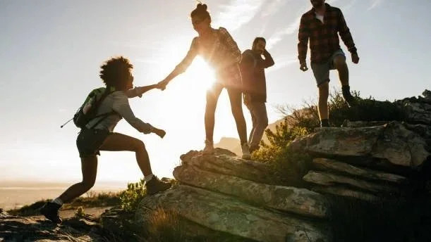 A group of four people hiking outdoors on rocky terrain during sunset, with one person helping another climb up a rock.