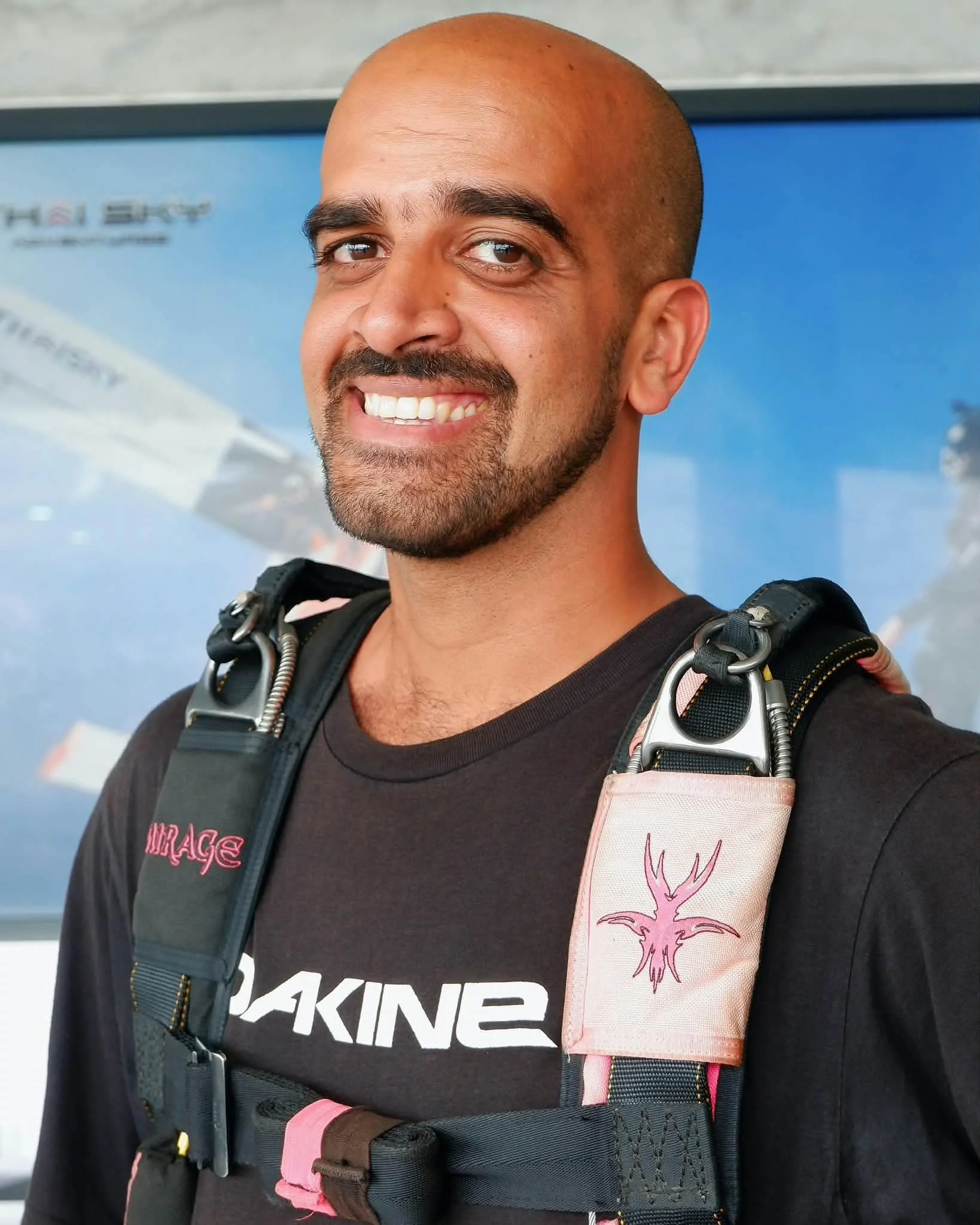 A man with a bald head, brown eyes, and a beard, smiling, wearing a black T-shirt with white and red text, a pink and black backpack, and a strap with a pink stick-on label featuring a pink deer antler illustration.