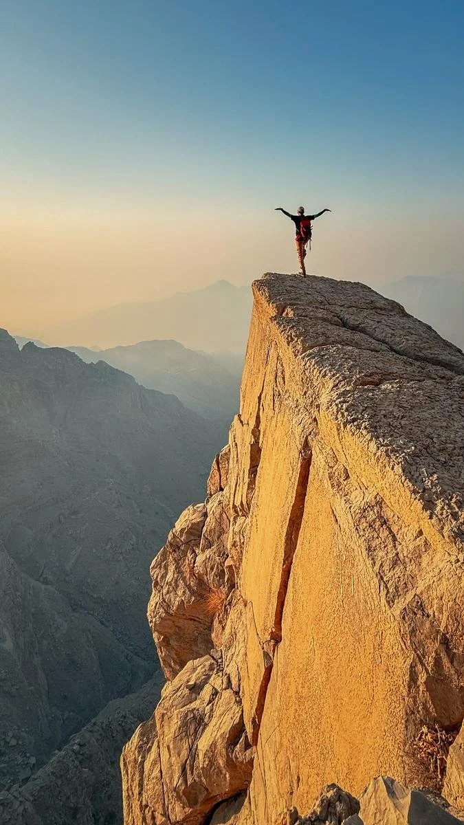 A person standing on the edge of a high, rocky cliff with arms outstretched, overlooking a mountain landscape at sunset or sunrise.