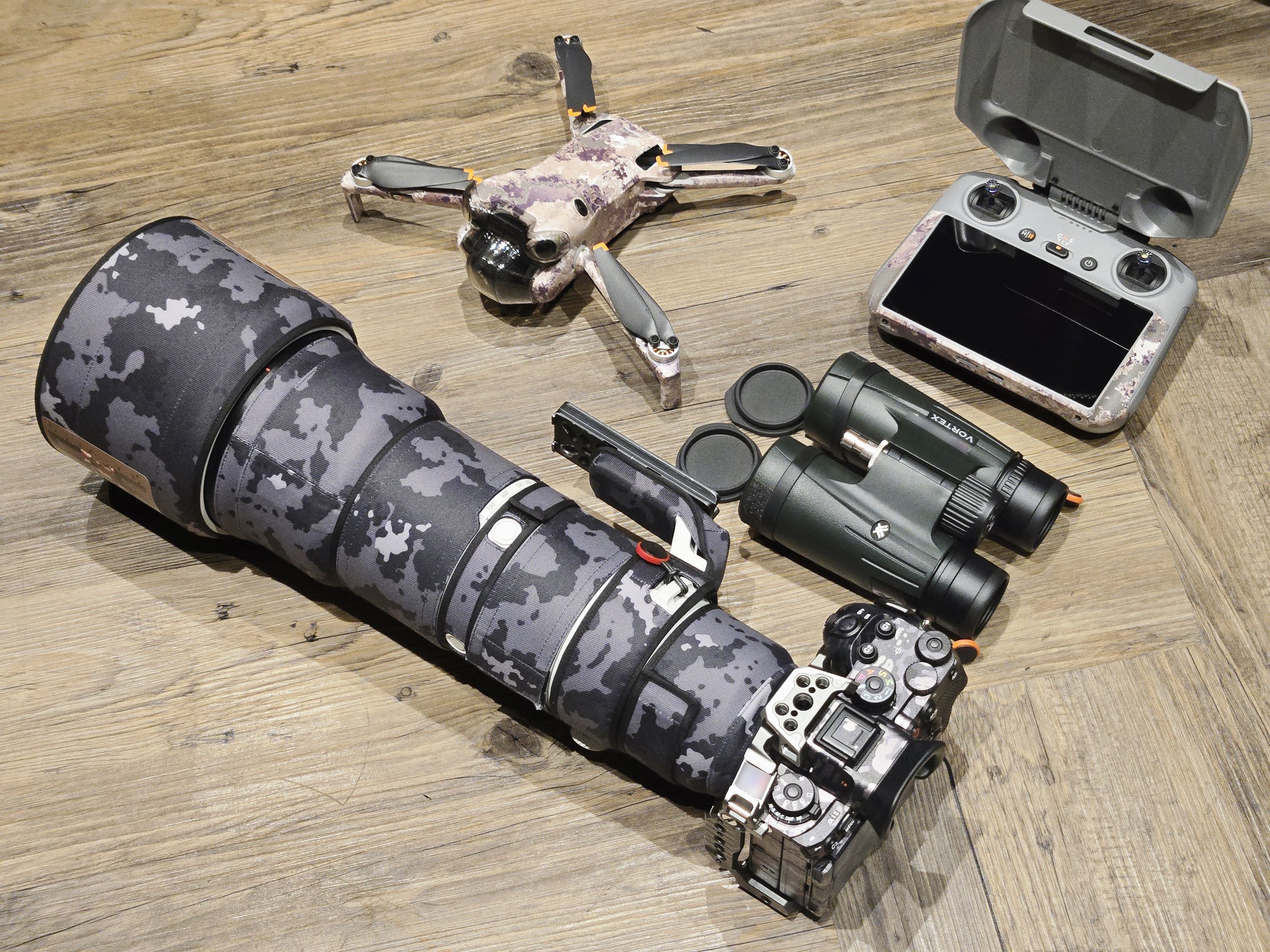 Camera equipment, a drone, binoculars, a remote control, and a camera on a wooden surface.
