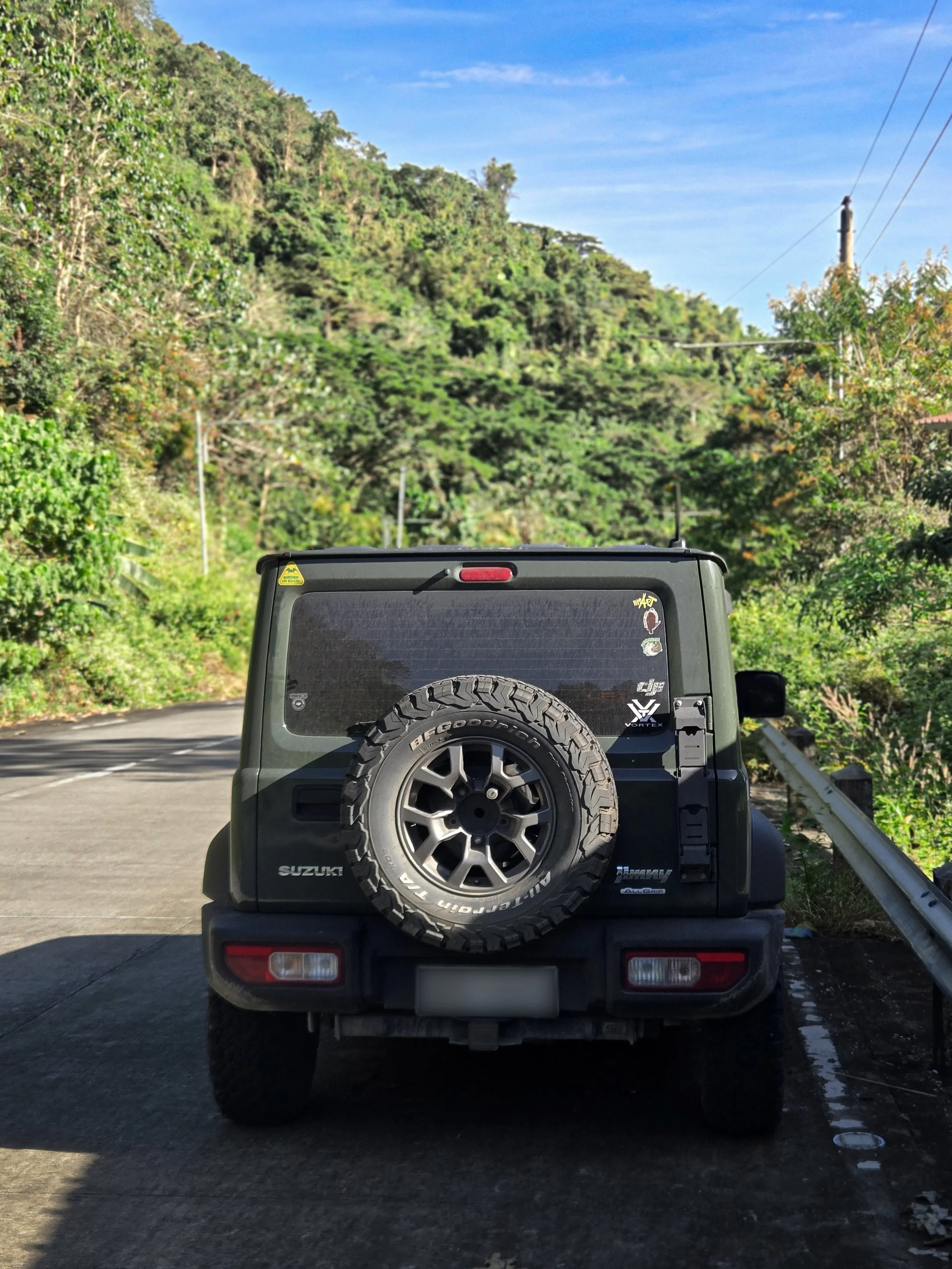 Rear view of a black Suzuki off-road vehicle with a spare tire mounted on the back, parked on the side of a winding road surrounded by lush green trees and hills under a clear blue sky.