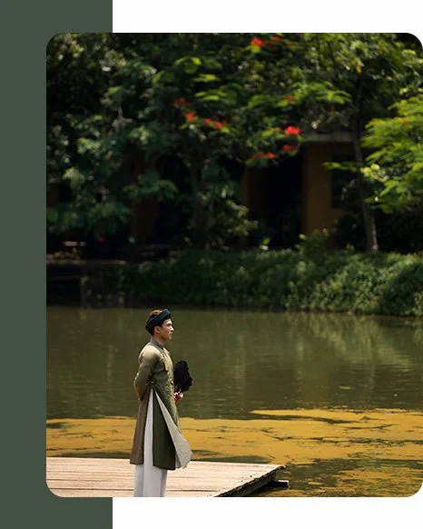 A woman in traditional clothing standing on a dock by a lake, surrounded by greenery and trees.