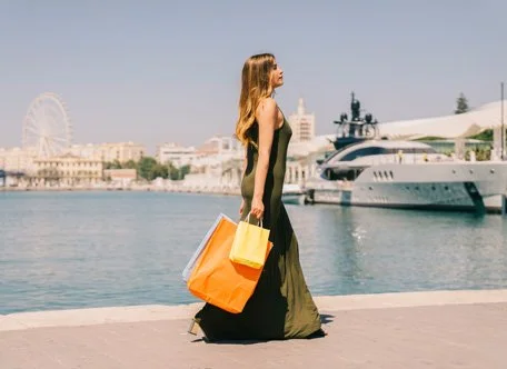 A woman in a black dress walking along a waterfront promenade with shopping bags, a ferris wheel, and boats in the background.