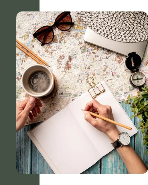 A top view of a workspace with a map, sunglasses, a cup of coffee, a notebook with blank pages, a pen, a gold paperclip, a wristwatch, a woven hat, a compass, and some greenery.