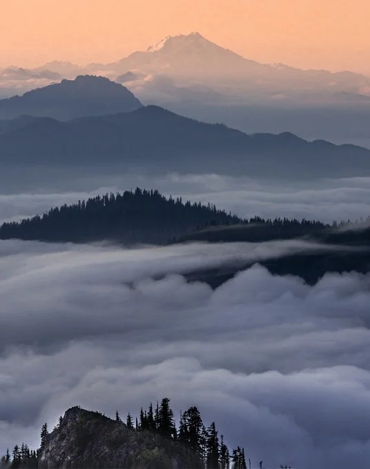 Layered mountain ranges with clouds in the valleys and a snow-capped peak in the background during sunrise or sunset.