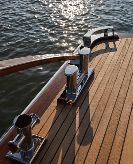View of a wooden dock with shiny metallic mooring cleats, a wooden handrail, and a body of water reflecting sunlight in the background.