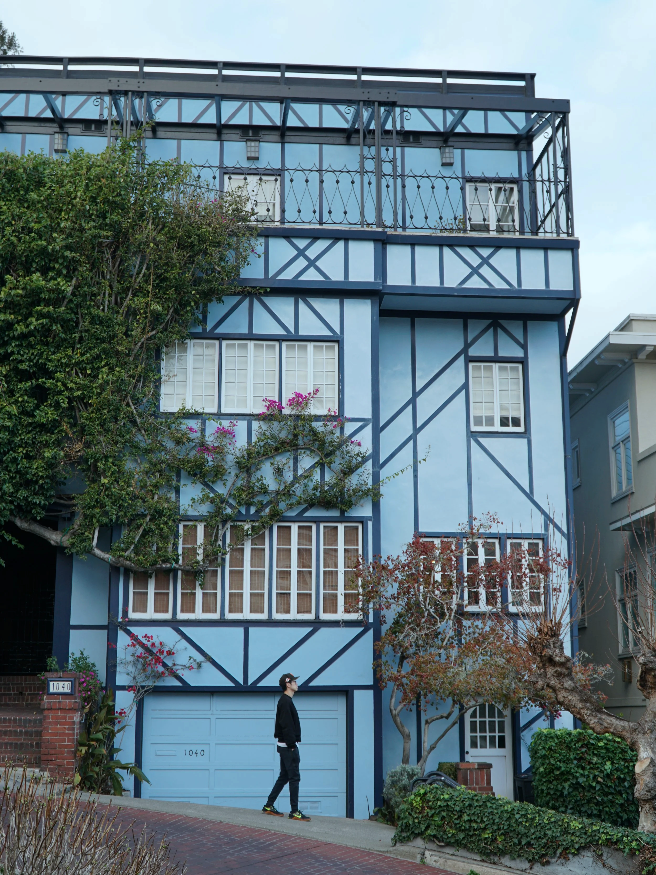 A person walking on the sidewalk in front of a multistory blue house with exposed dark blue wall framing, white windows, and small trees and bushes in the yard.