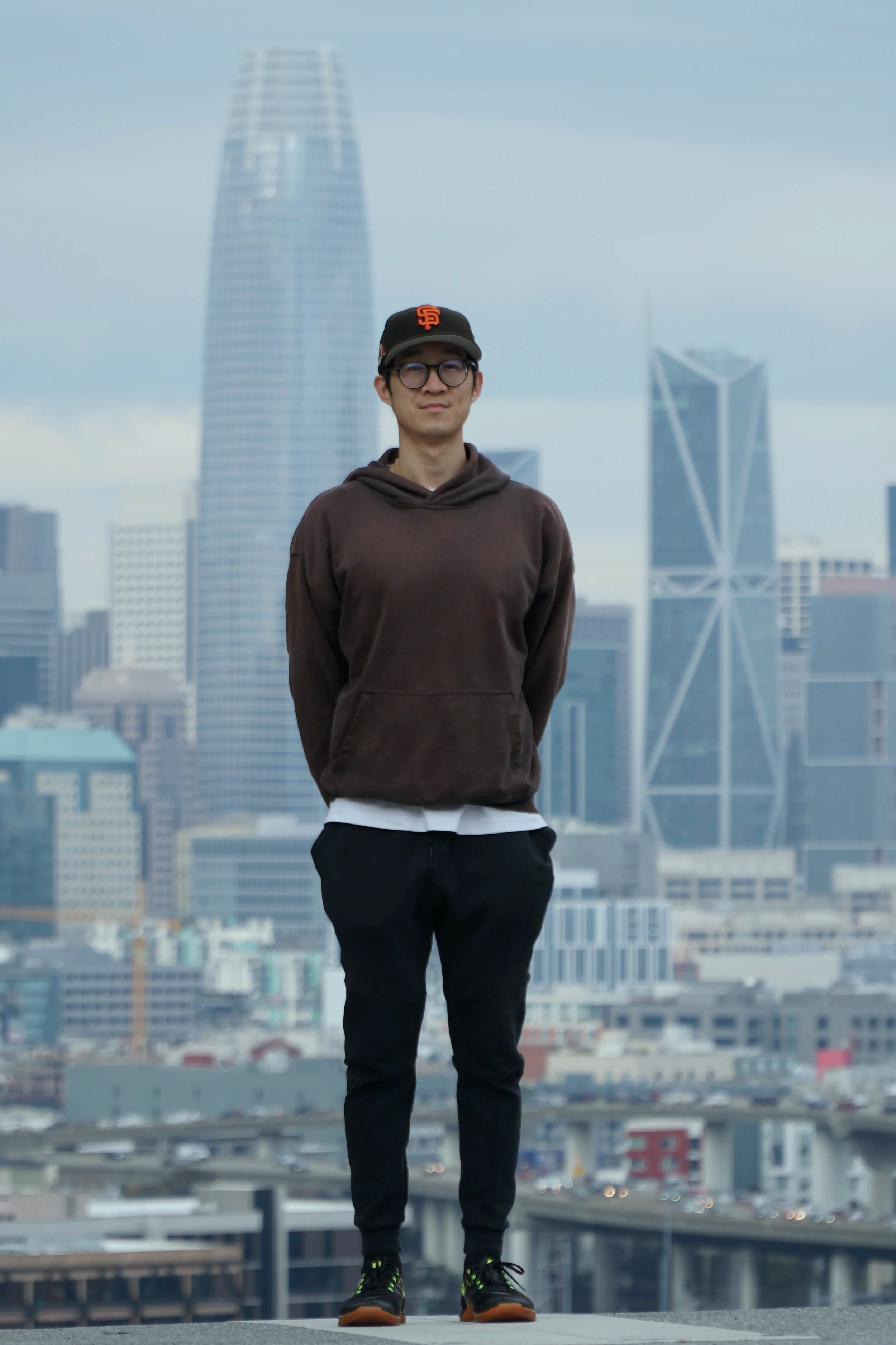 A young man wearing glasses, a black San Francisco Giants cap, a brown hoodie, black pants, and black sneakers standing outdoors on a rooftop with a city skyline in the background.
