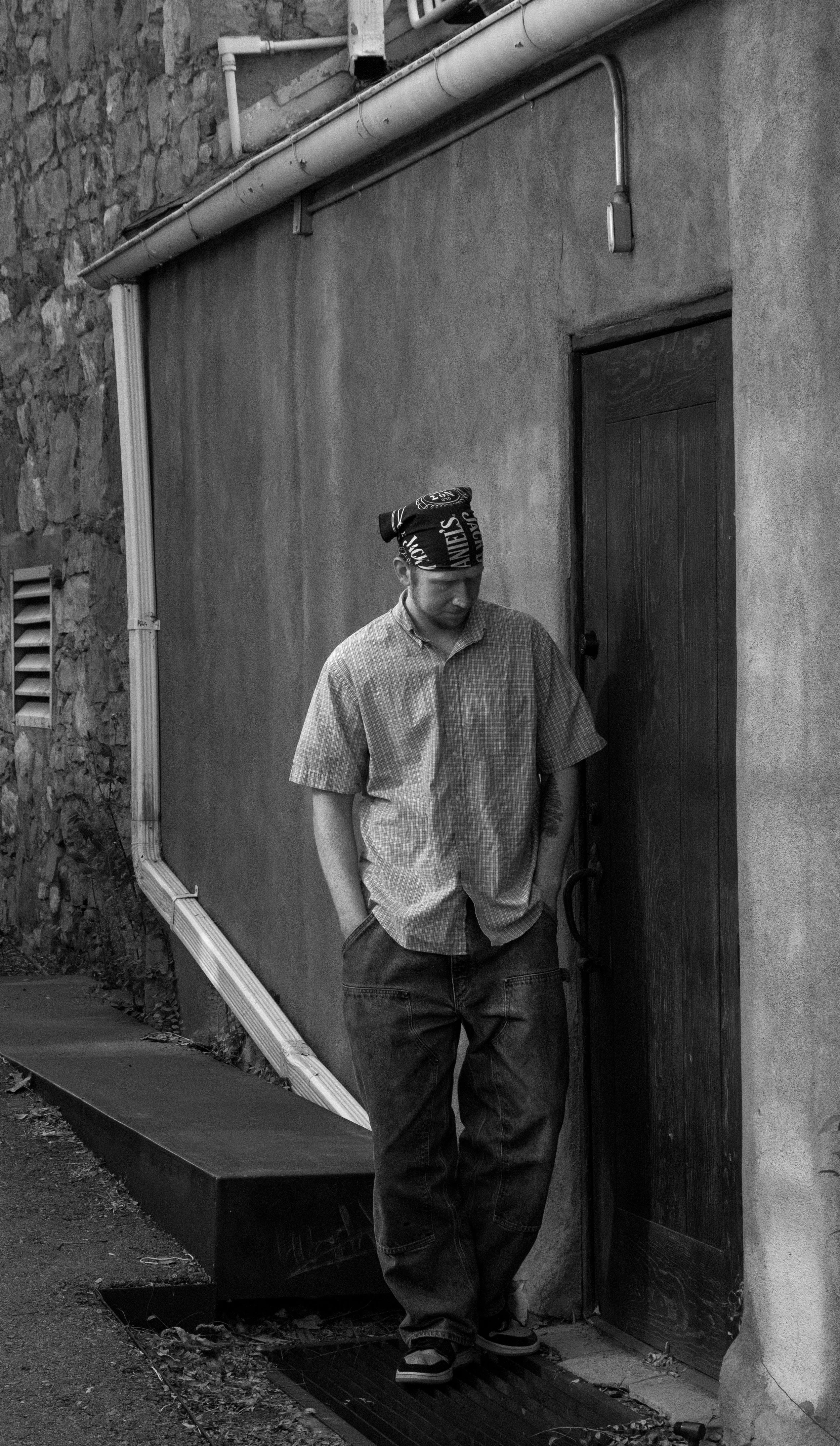 A man standing outdoors in front of a door, wearing a checkered shirt, baggy jeans, and a bandana on his head, with his hands in his pockets.
