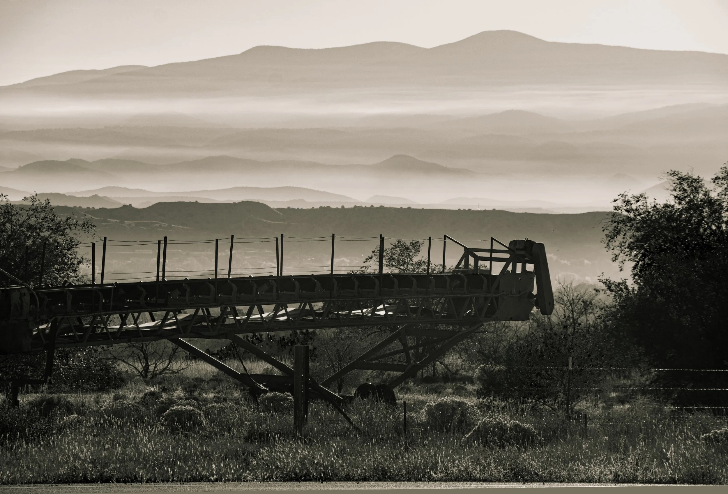 Black and white photo of an old tractor or farming equipment in a field with trees and hills in the background, misty mountains in the distance.