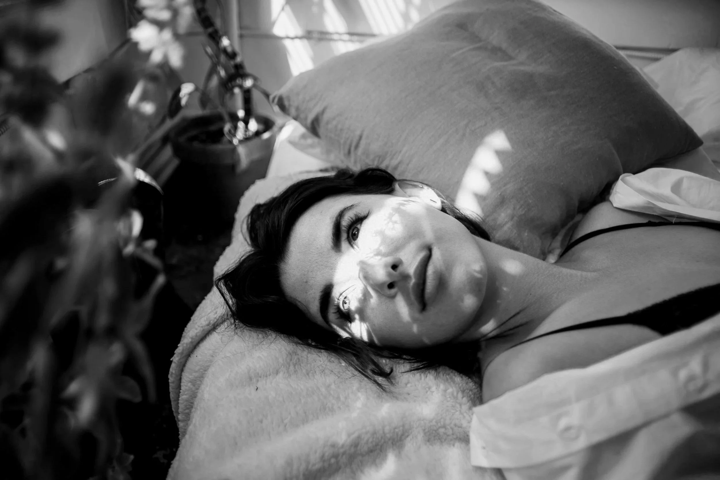 A woman with dark hair lying on her back on a bed, with light and shadow patterns on her face from window blinds, in a black and white photo.