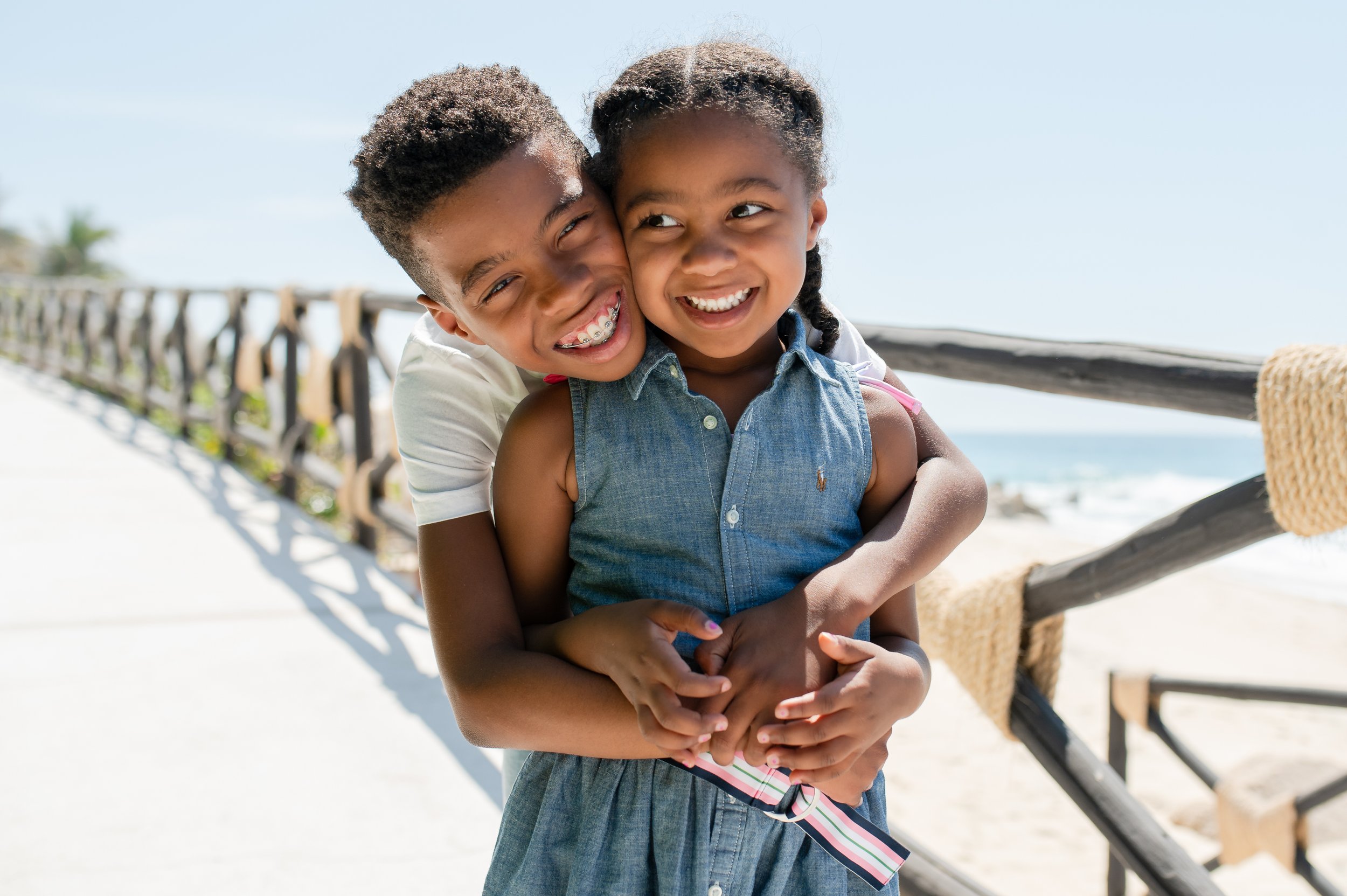 Dos niños sonreíntiendo y abrazándose en una playa debajo de un arco de madera, con mar y cielo despejado al fondo.