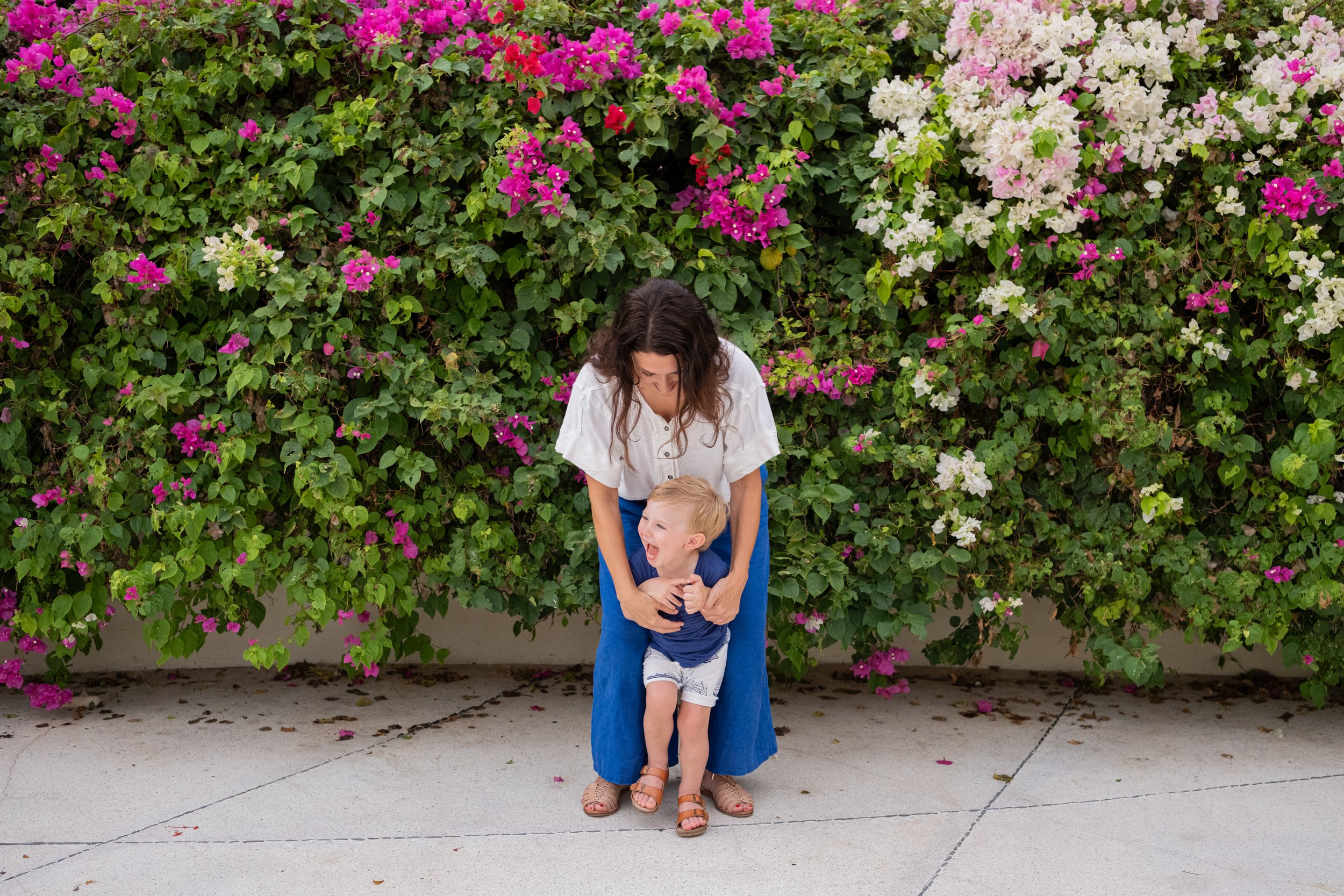 Una mujer y un niño pequeños riendo y jugando frente a un arbusto de flores de colores rosados, blancos y violetas.