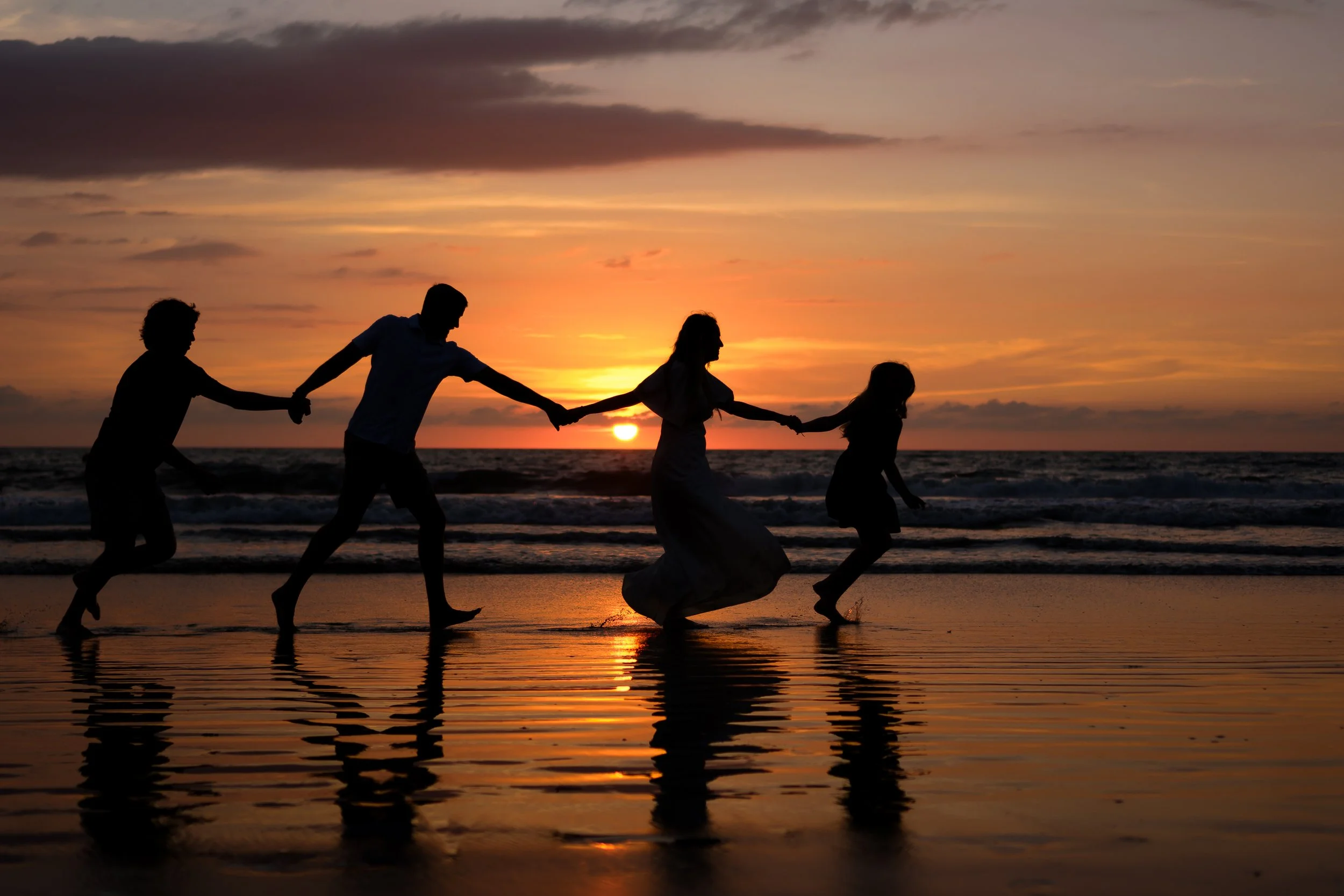 Una familia de cinco personas formando una cadena con las manos en la orilla del mar durante un atardecer, con el sol ocultándose en el horizonte y reflejándose en el agua.