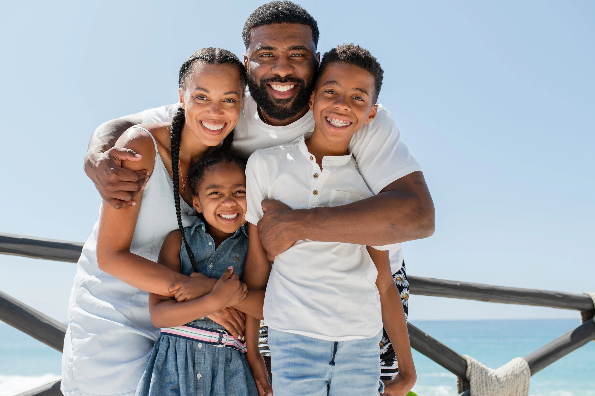 Familia sonriente en la playa abrazándose y posando para la foto bajo un cielo azul.