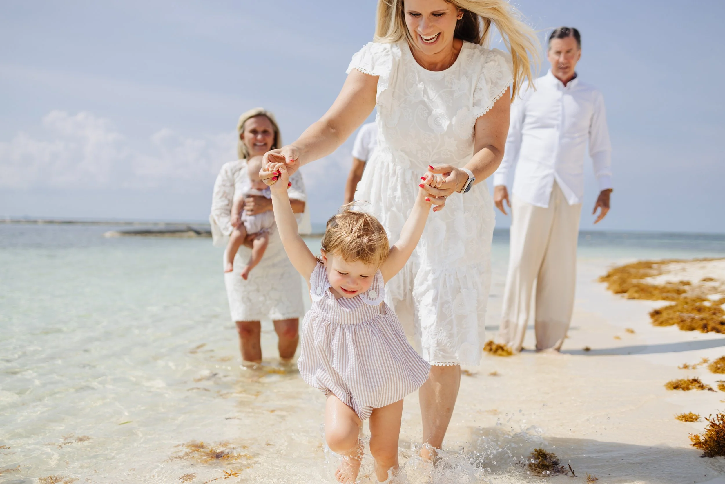 Un grupo de personas en la playa, disfrutando y jugando con un niño pequeño que está aprendiendo a caminar en el agua, acompañados por mujeres y un hombre, con mar y cielo azul de fondo.