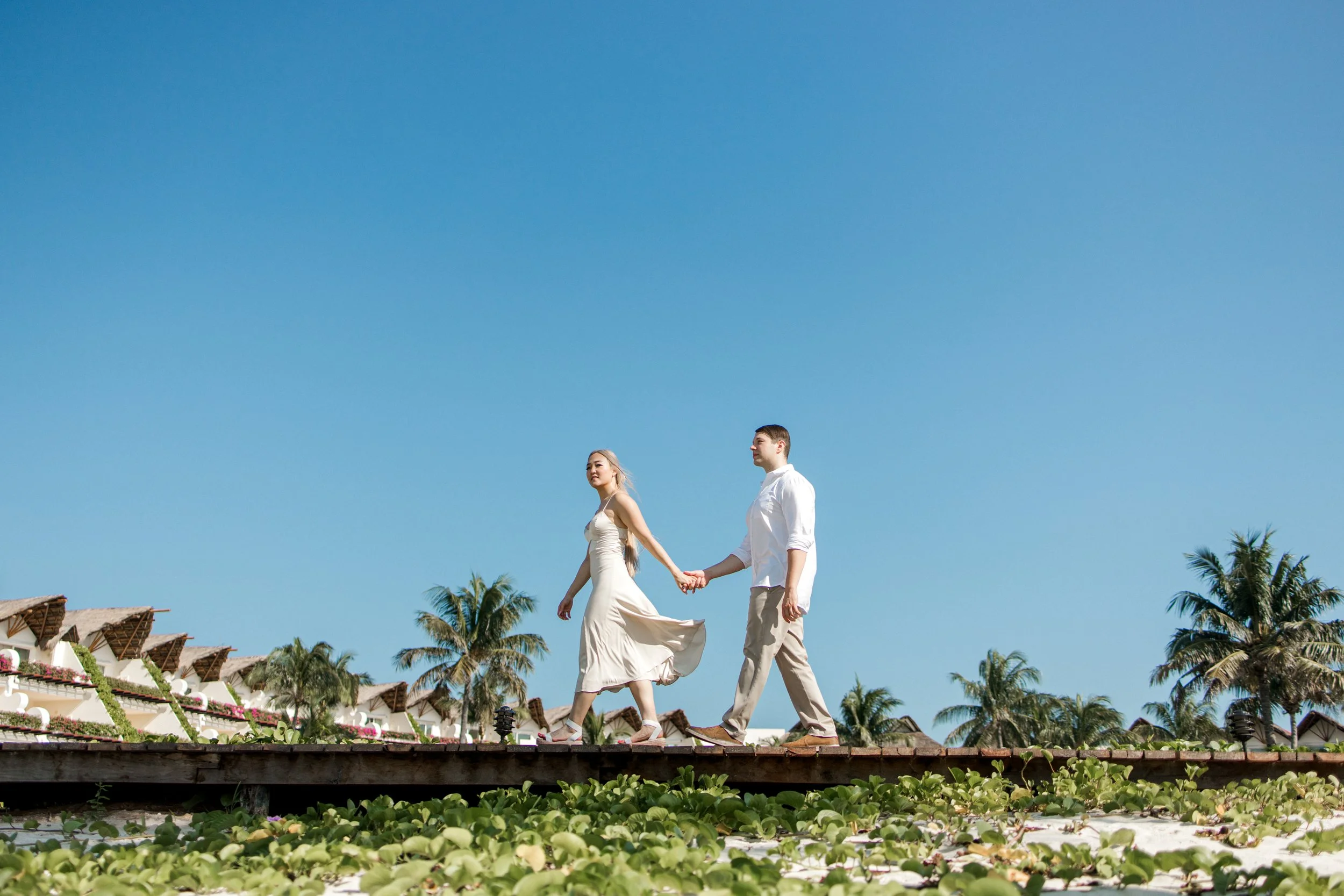 Pareja caminando de la mano en una playa con palmeras y casas de estilo tropical bajo un cielo despejado.