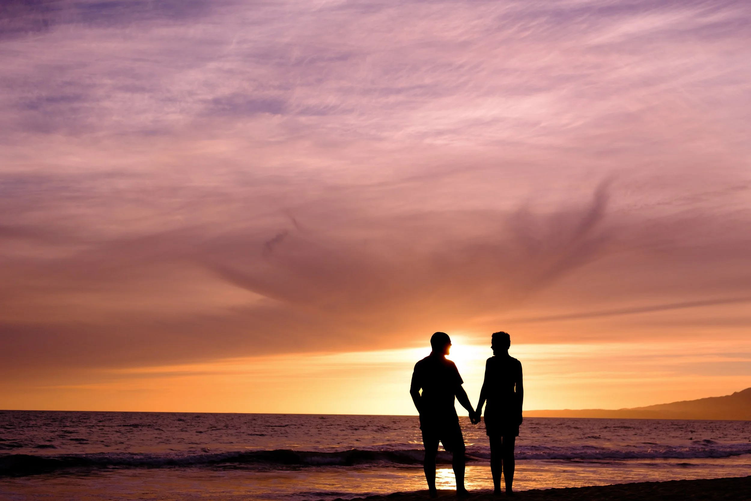Una pareja de personas de perfil, de pie en la playa, sosteniéndose de la mano durante un atardecer con cielo colorido y mar en el fondo.