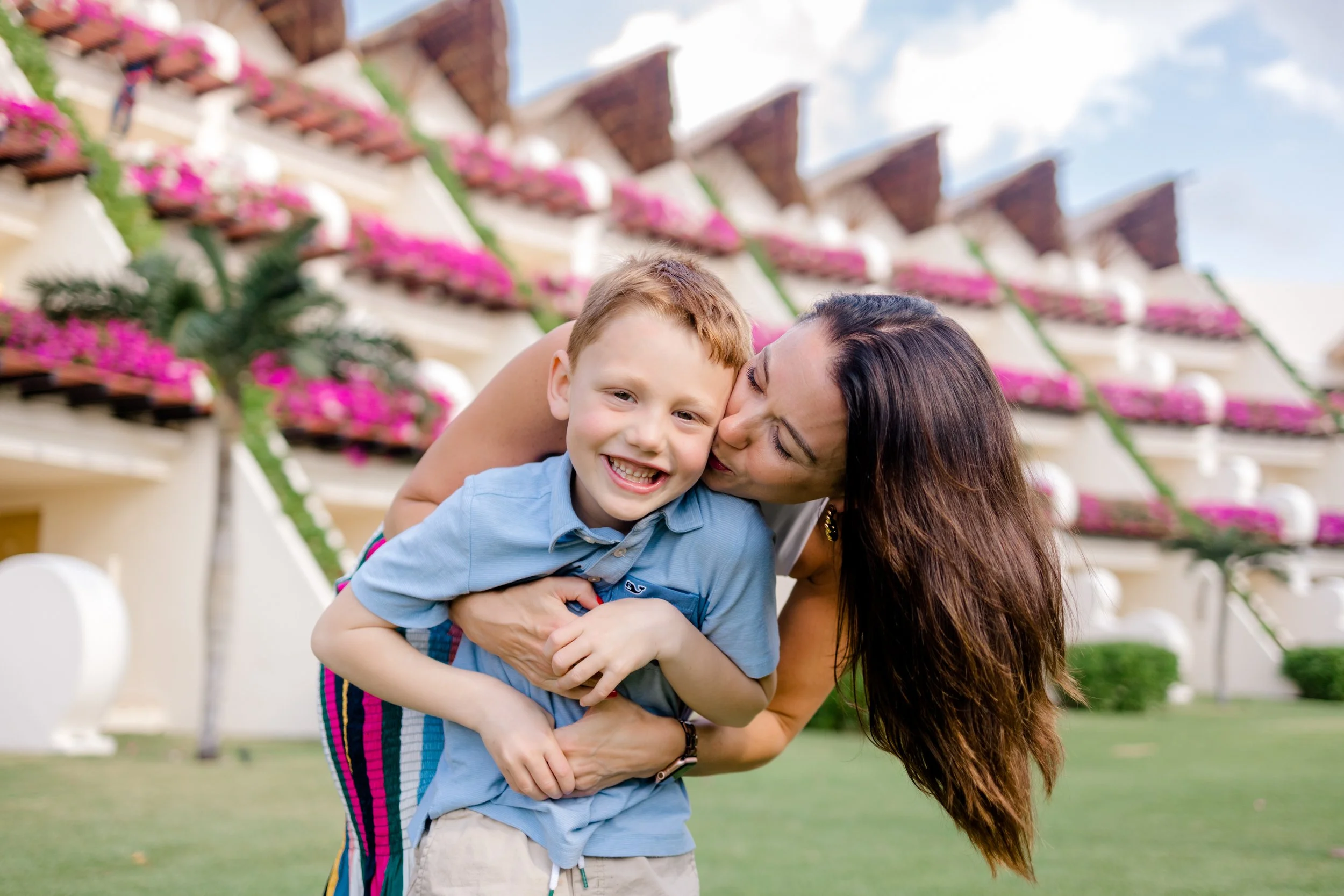 Una mujer y un niño disfrutando un momento de alegría al aire libre frente a un edificio con balcones llenos de flores rosadas.