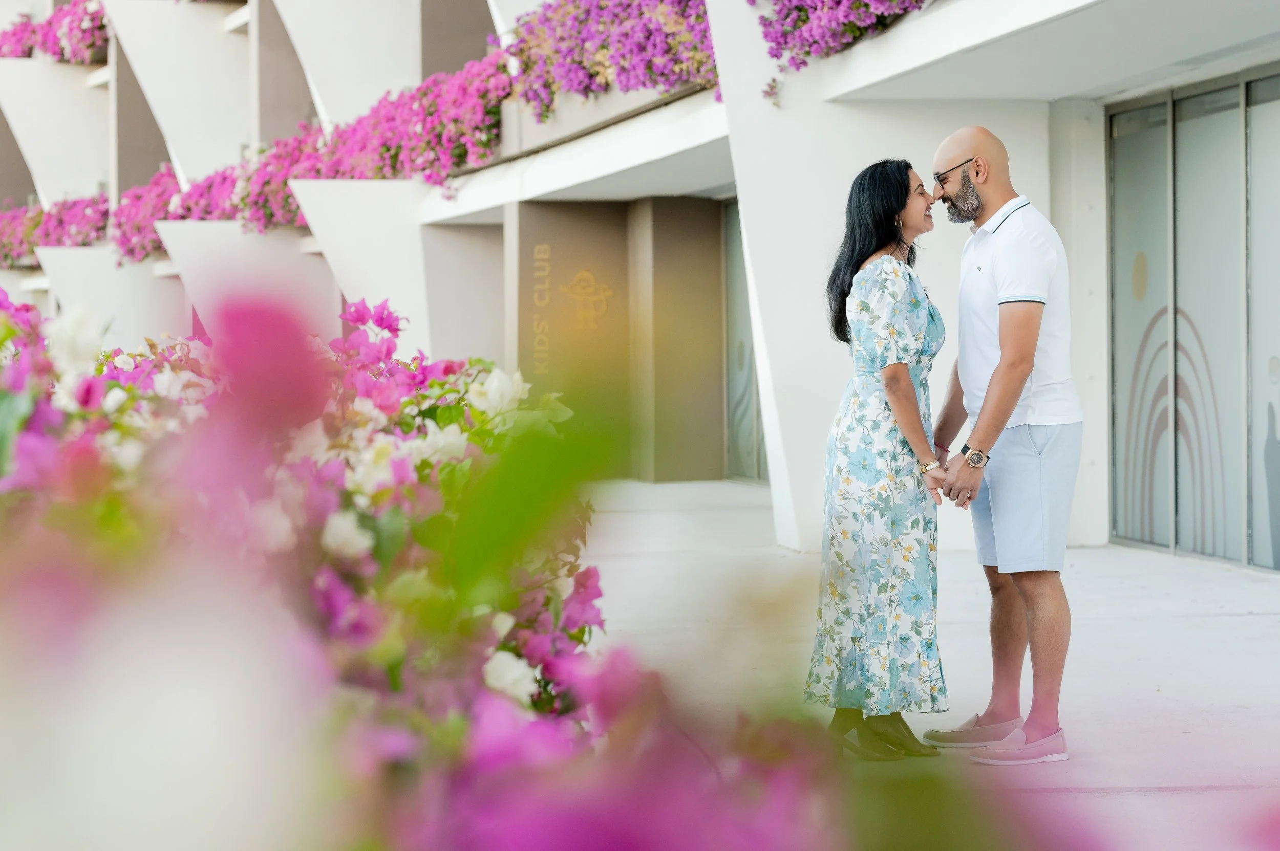 Una pareja de hombres y mujer con vestimenta veraniega se mira cariñosamente, tocándose las narices, en un espacio con muchas flores rosas en el fondo.