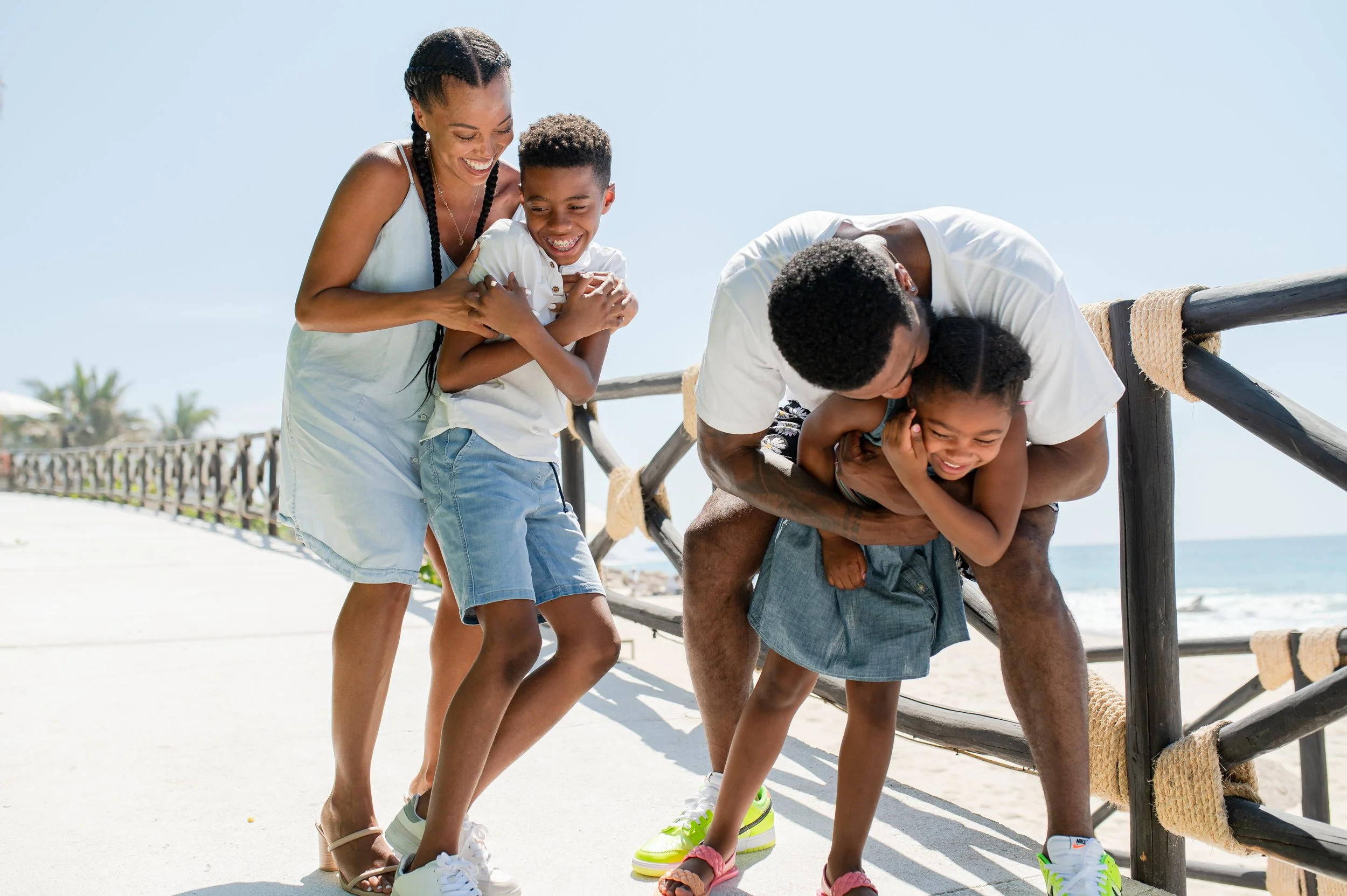 Familia disfrutando en la playa, riendo y jugando juntos con dos niños y dos adultos, al lado de una cerca de madera en un día soleado.