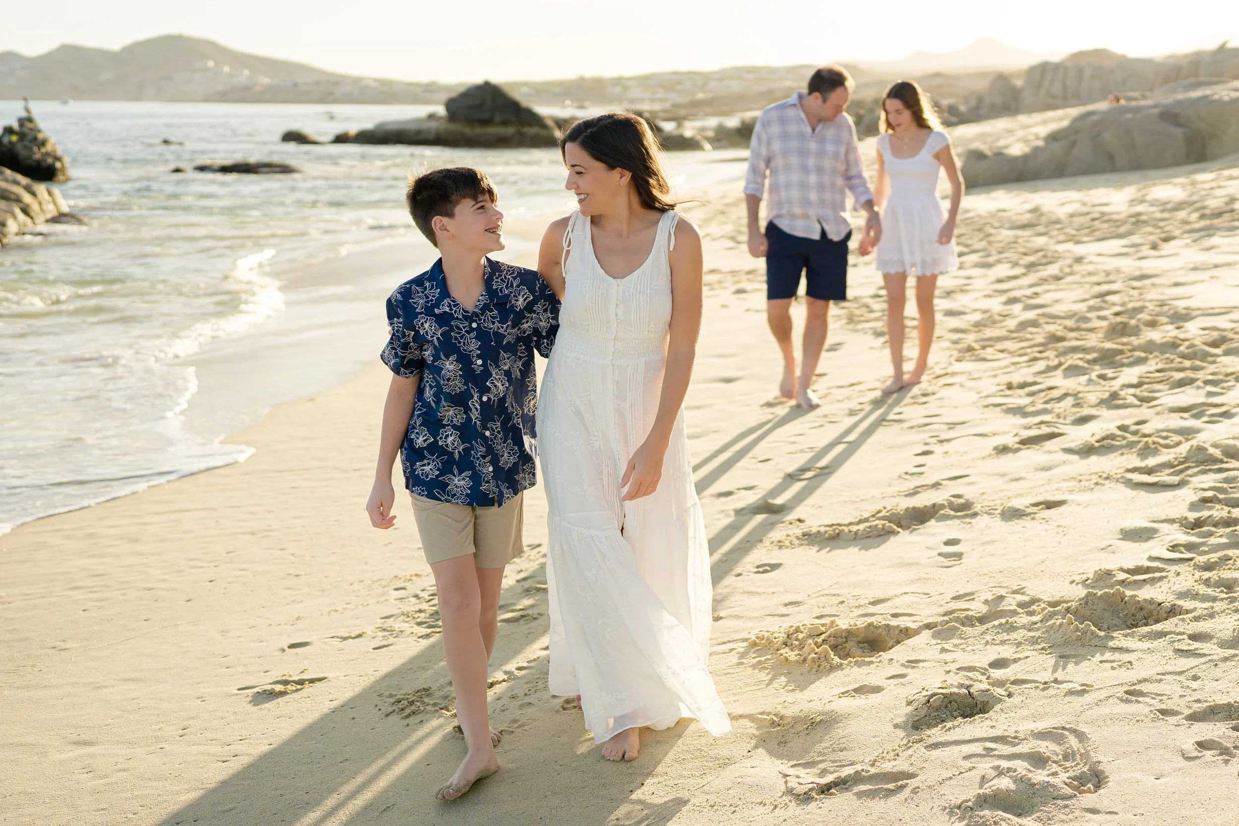 Grupo de cinco personas caminando en la playa, sonriendo y disfrutando del momento, con el mar y rocas al fondo.
